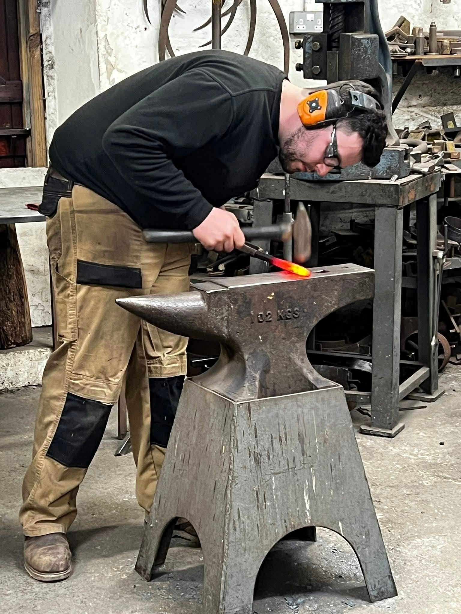 A man in protective gear forging hot metal on an anvil in a workshop.