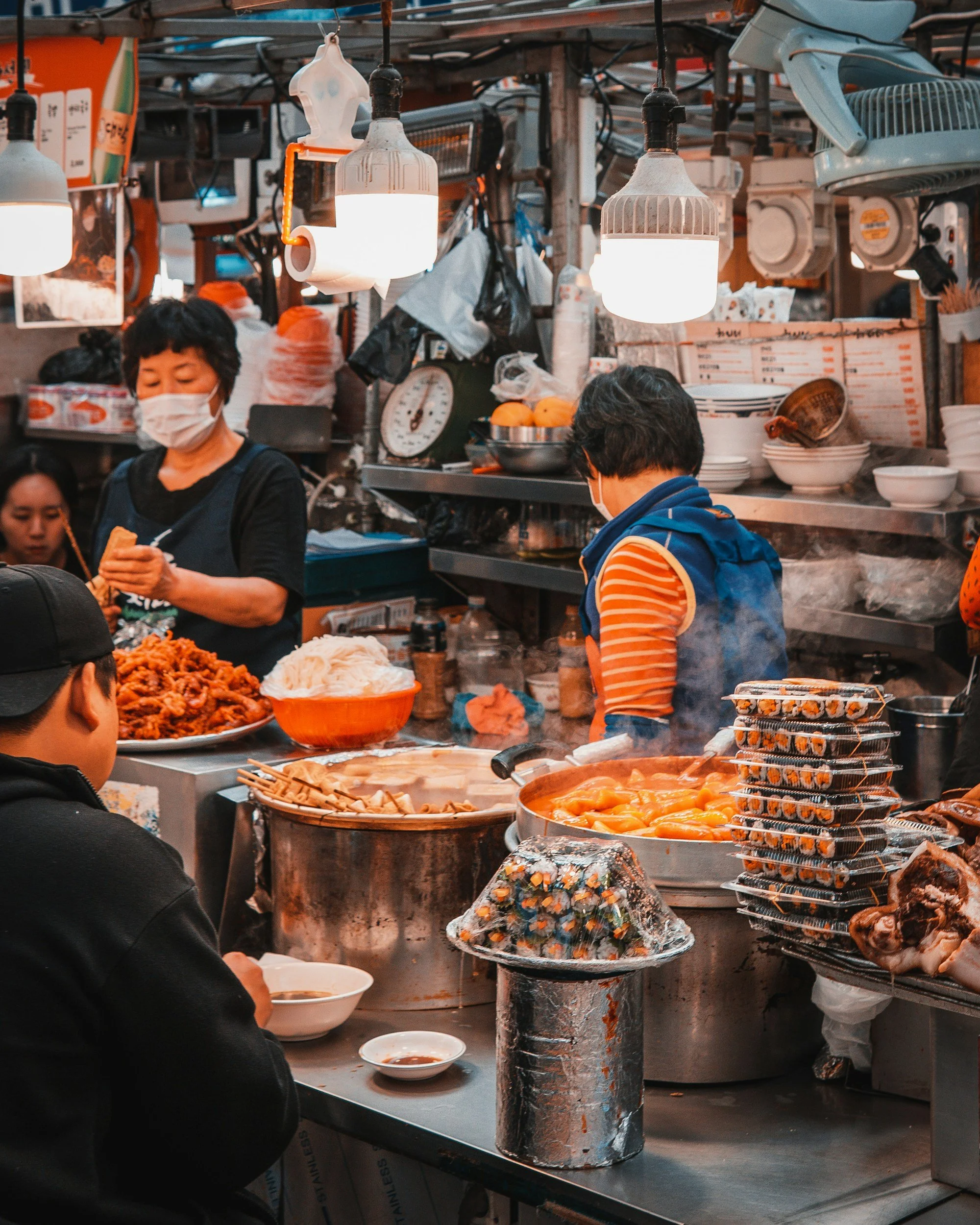 A street food stall with hanging lamps, various dishes including kimchi and noodles, containers, a woman wearing a face mask preparing food, and a person in a striped orange and white shirt with a blue vest eating.