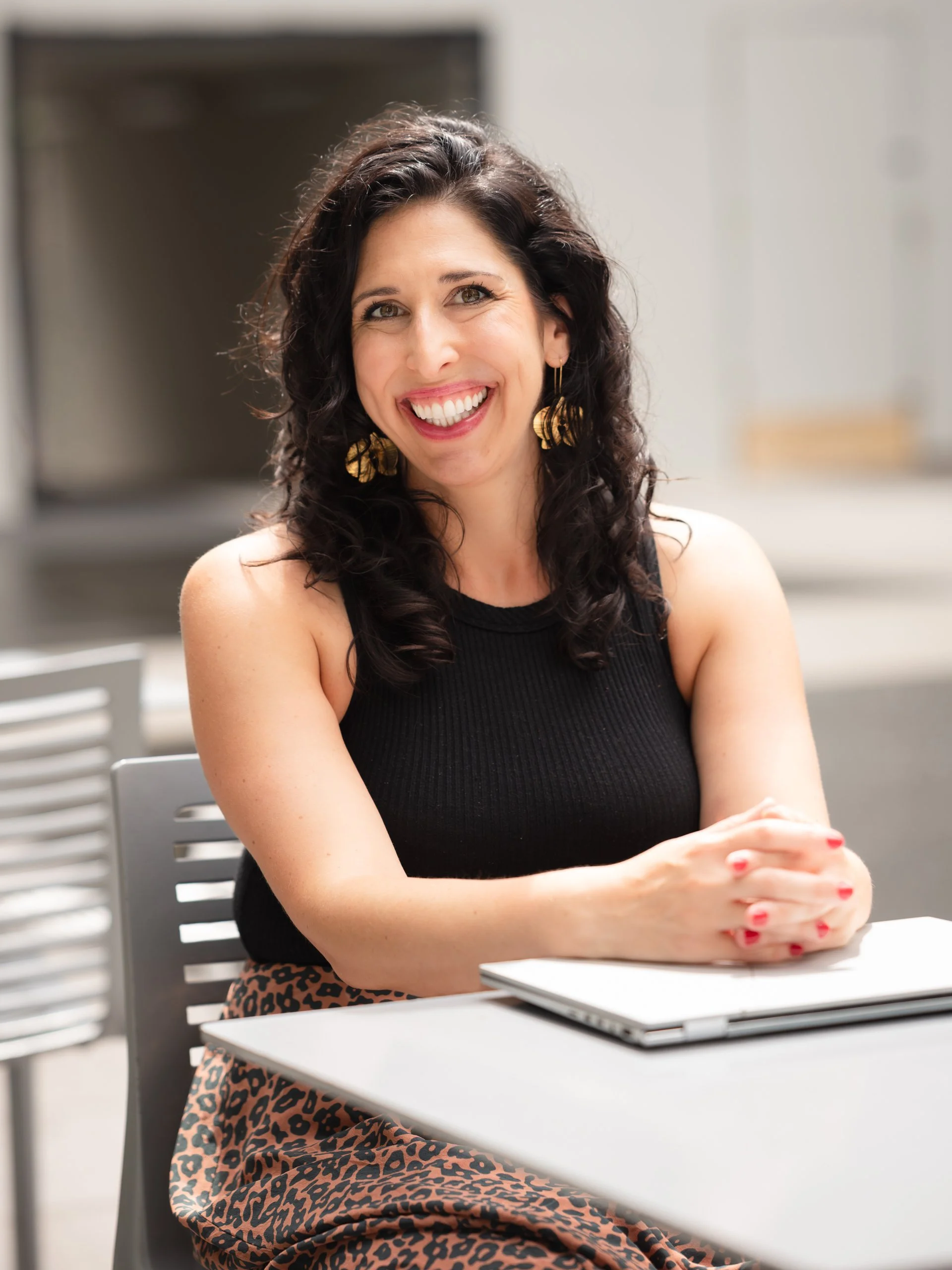 A woman with dark curly hair smiling at the camera, sitting at a table with a laptop, wearing a black sleeveless top, gold earrings, and a leopard print skirt.