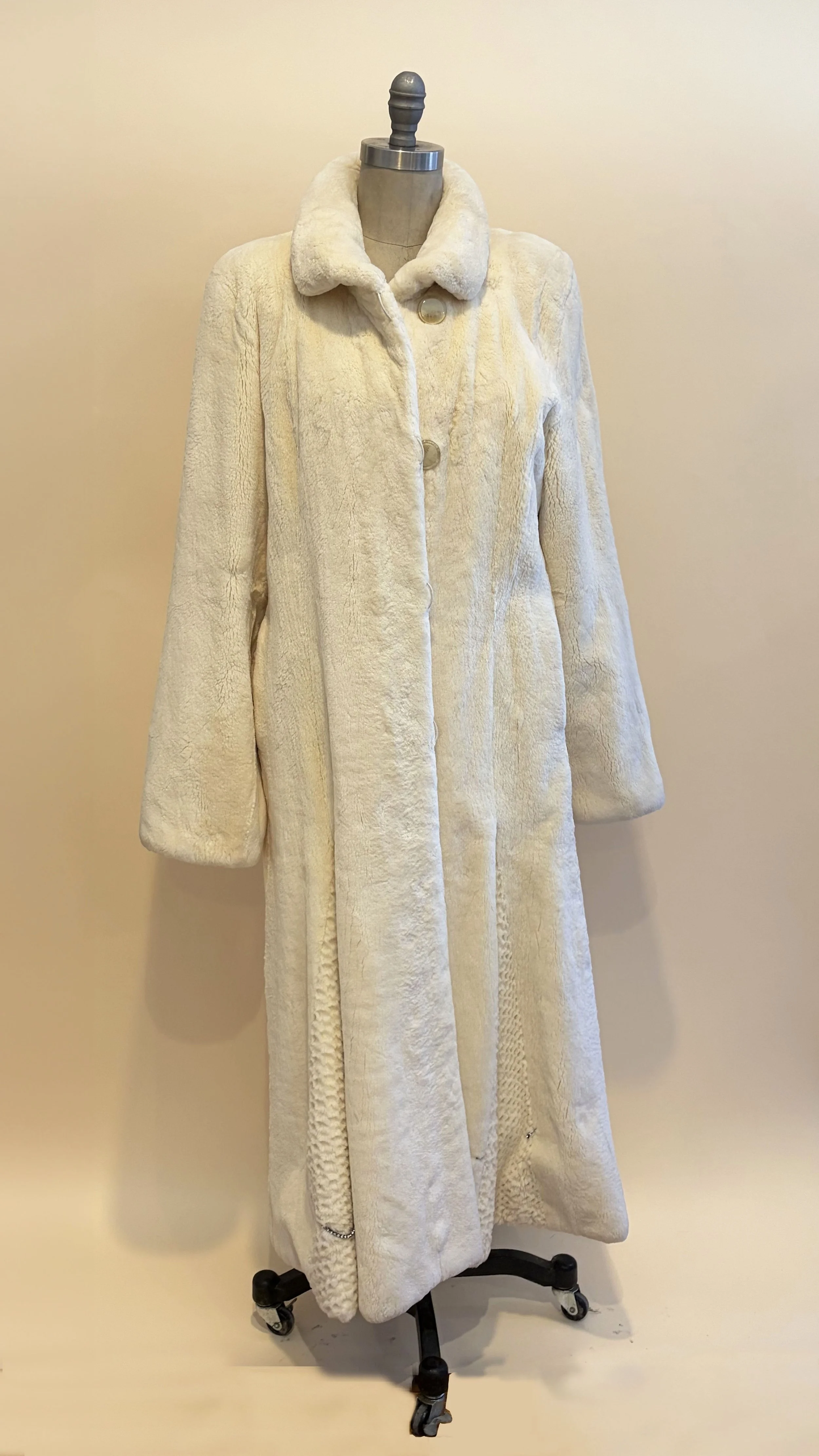 A long, cream-colored, plush coat on a mannequin with a metal stand on wheels against a plain beige wall.
