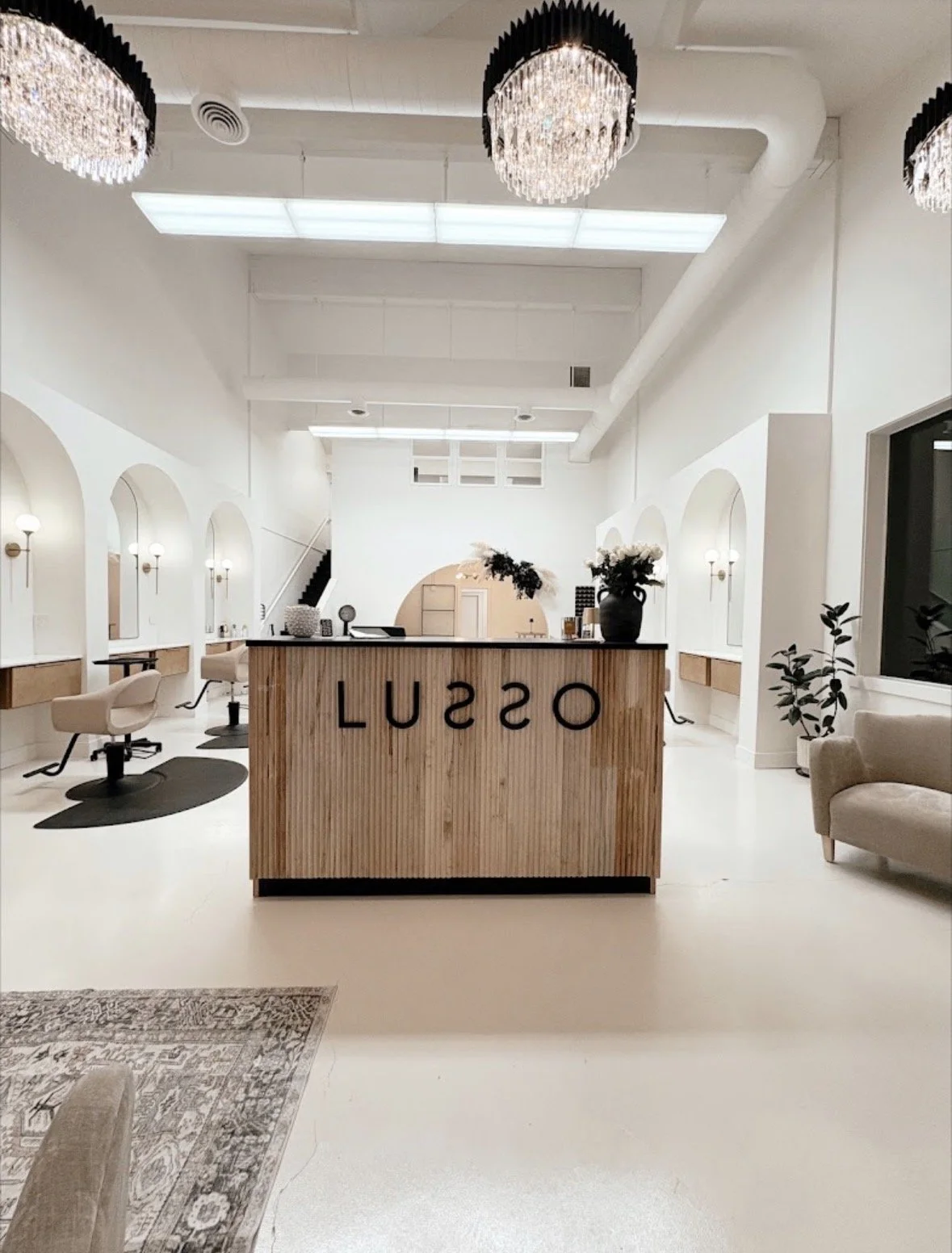 Interior of a modern hair salon with white walls, ceiling, and furniture. The reception desk is made of light wood with the word 'OSSU' written backward. There are chairs for clients, mirrors along the walls, and elegant chandeliers hanging from the 