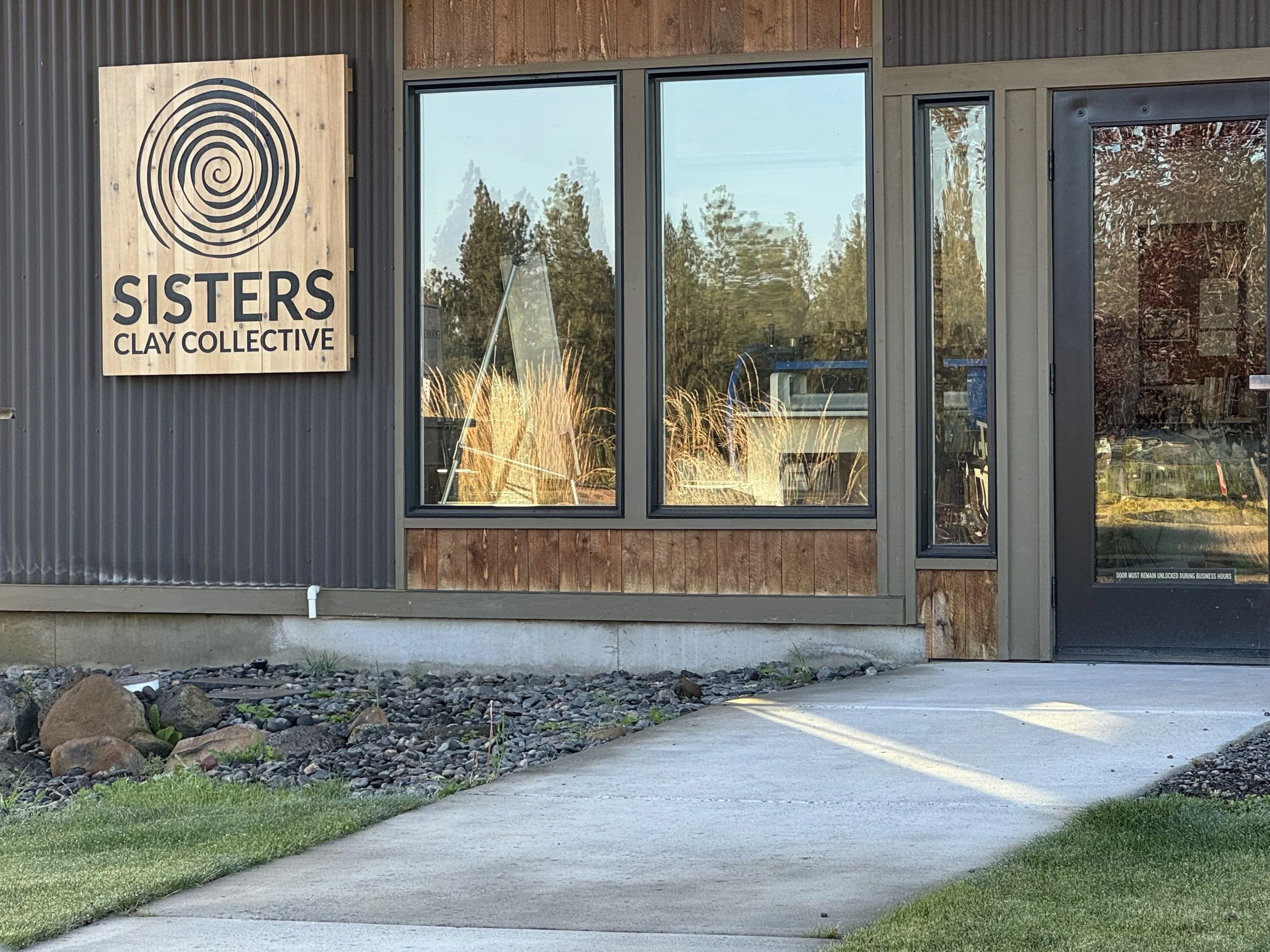 Exterior of a building with wooden and metal siding, large glass windows, and a sign that reads 'SISTERS CLAY COLLECTIVE' with a spiral logo. The building is next to a cement sidewalk with a grassy area and rocks in the foreground.