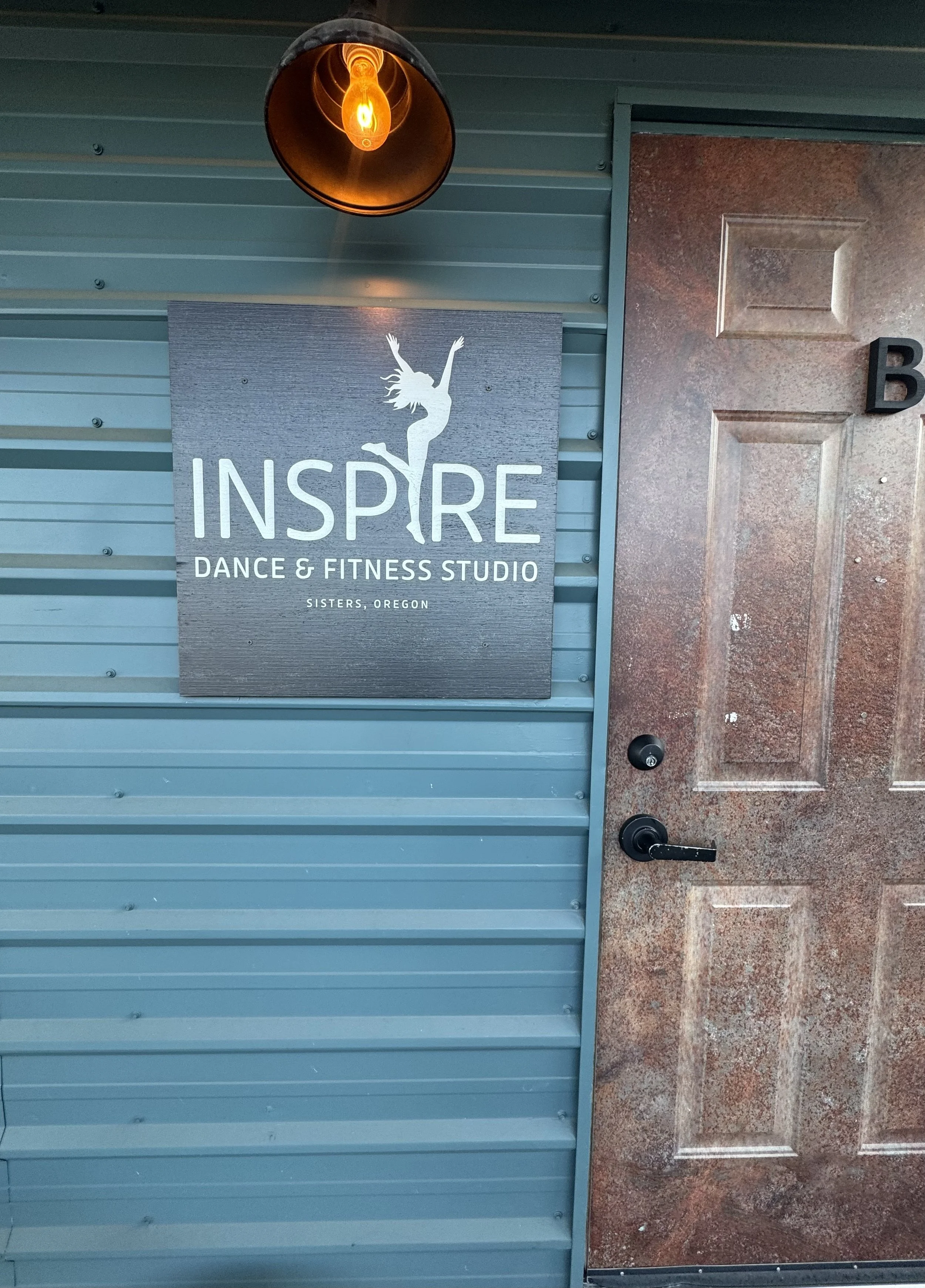 Blue exterior wall with a sign that reads 'Inspire Dance & Fitness Studio, Sisters, Oregon,' featuring a silhouette of a woman dancing, with overhead lighting and a brown tiled floor.