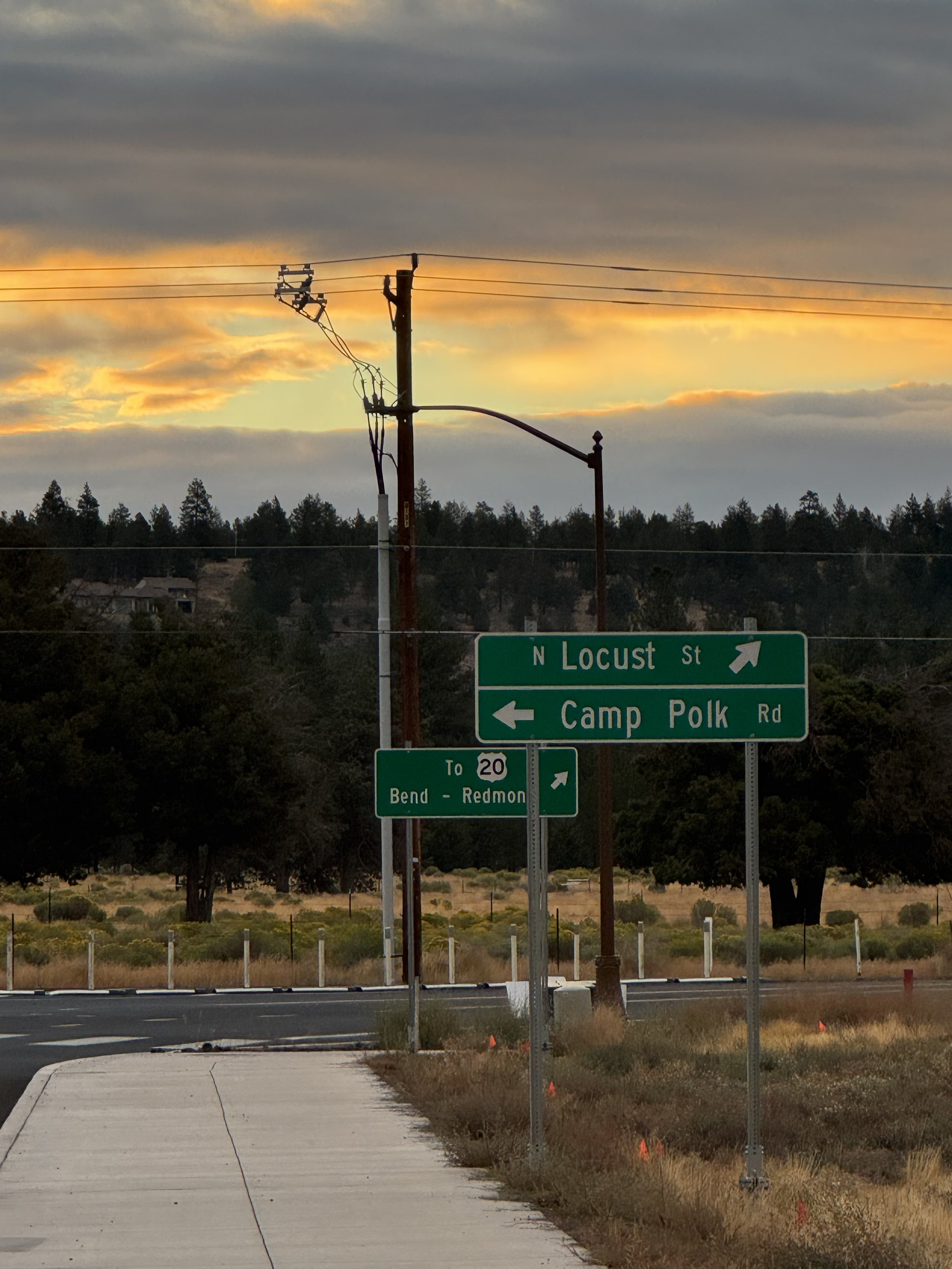 Street scene at sunset with road signs pointing to North Locust Street and Camp Polk Road, power pole with power lines, trees in background, and a partly cloudy sky with orange and gray clouds.