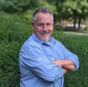 A man with glasses, gray hair, and a beard, smiling with arms crossed, standing outdoors with green bushes and trees in the background.