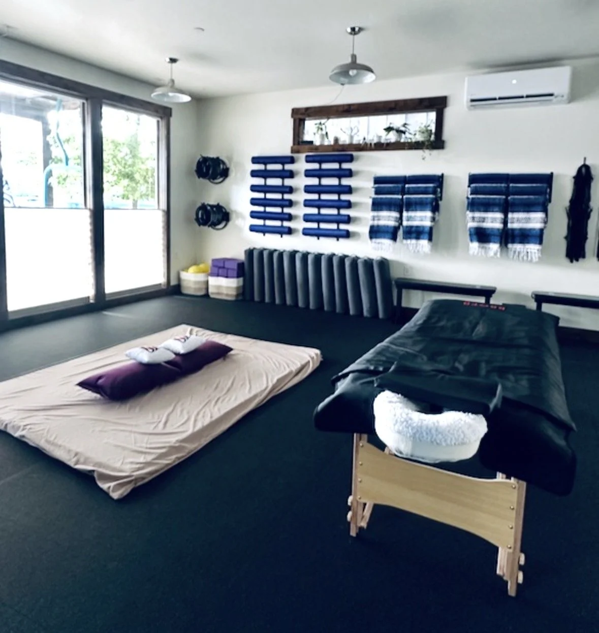 A room with flooring mats, pillows, and bedding, featuring an assortment of rolled blue mats on the wall, towels, and a massage or therapy table.