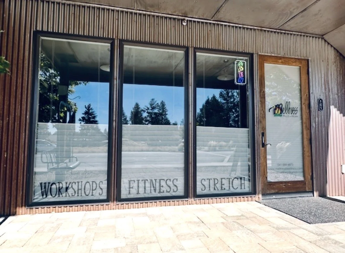  storefront window with words 'WORKSHOPS,' 'FITNESS,' and 'STRETCH' written on the lower part of the glass, and a glass door with a logo and contact information, wooden framing, and an outdoor sidewalk.