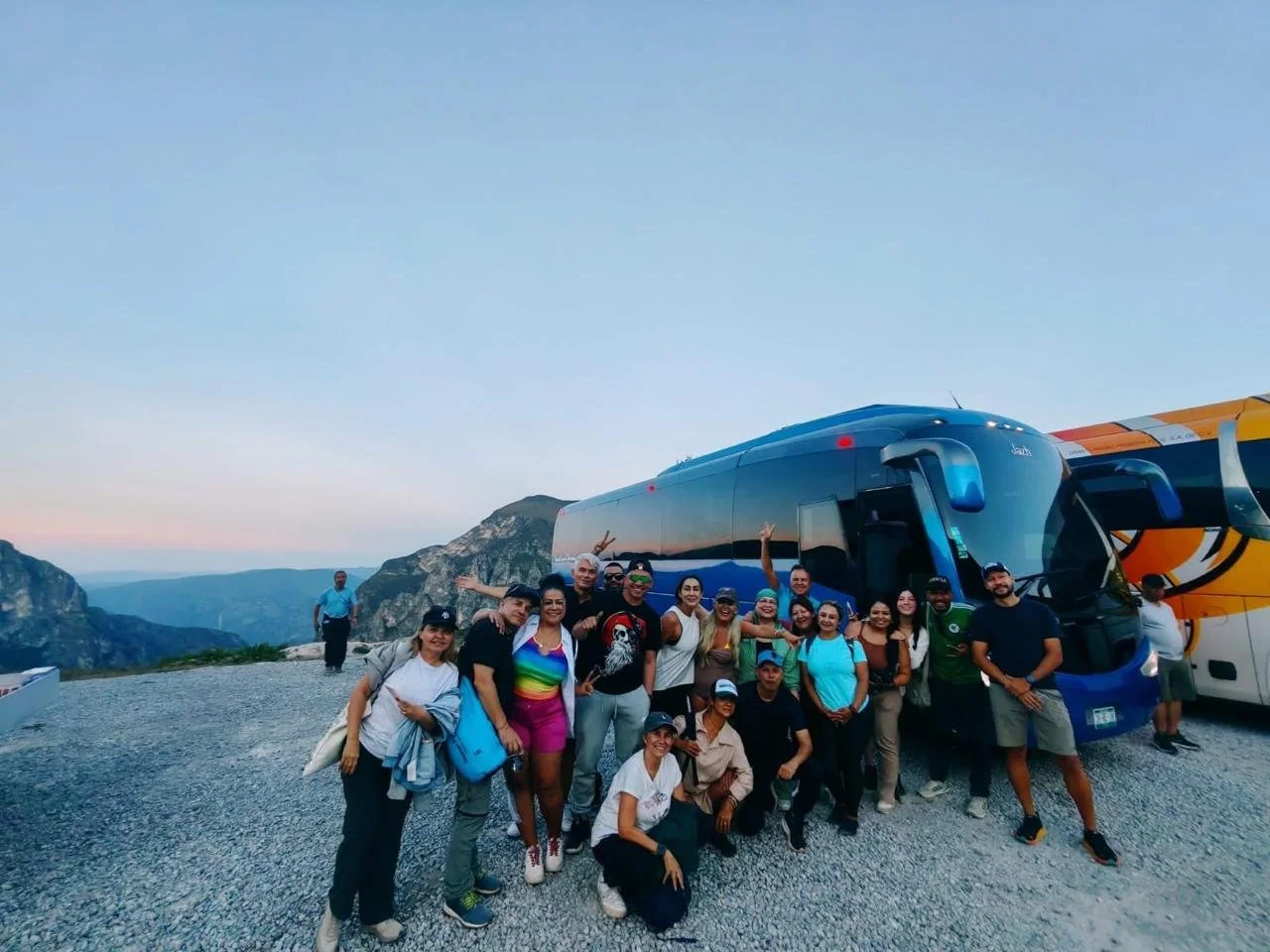 Grupo de personas sonriendo y haciendo gestos de paz frente a autobuses en un paisajes montañoso al atardecer.