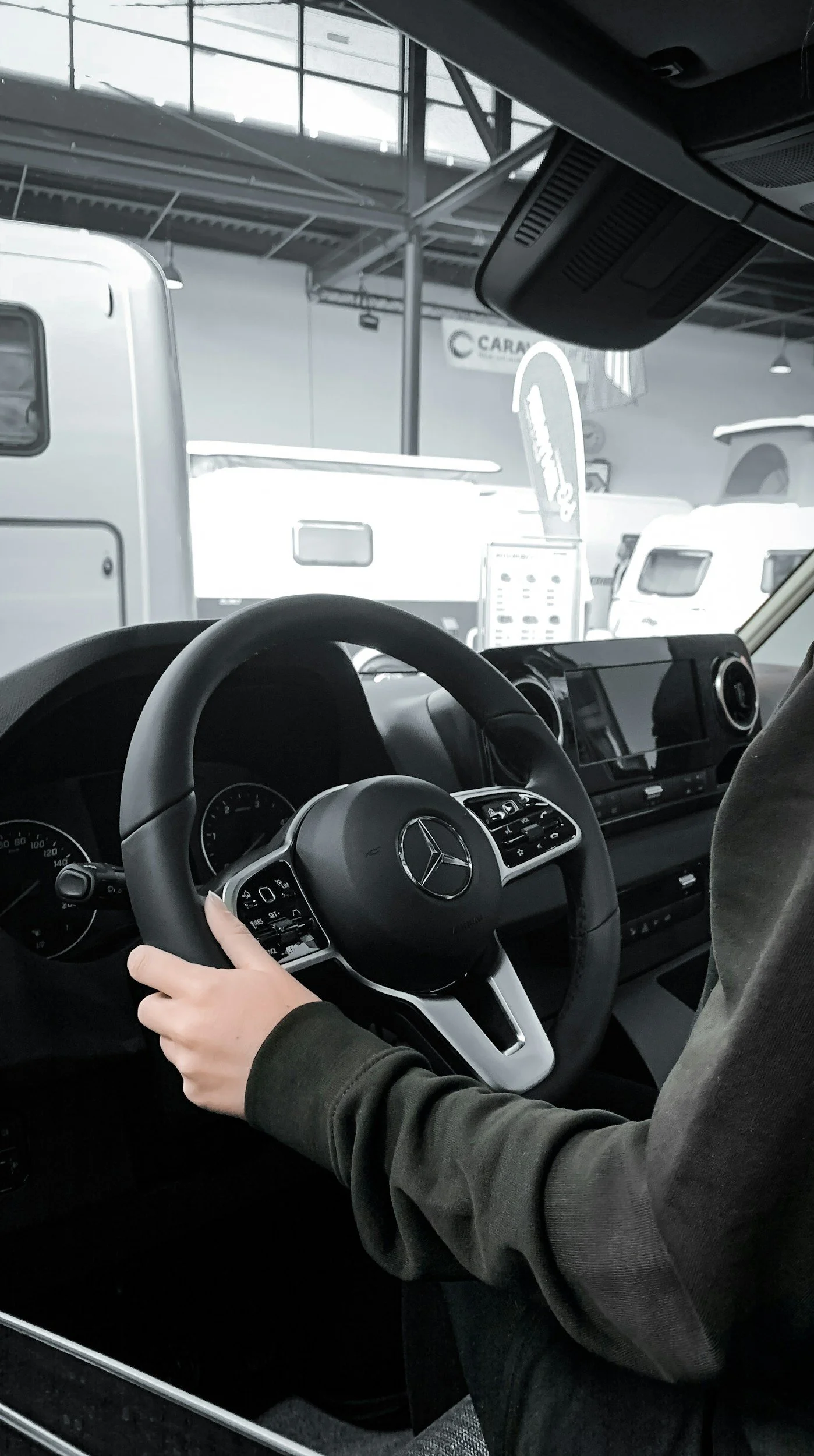 Interior de un coche Mercedes-Benz con una persona al volante, mano en el volante, en un lugar que parece ser un concesionario de autos con otros vehículos y señalización visible.
