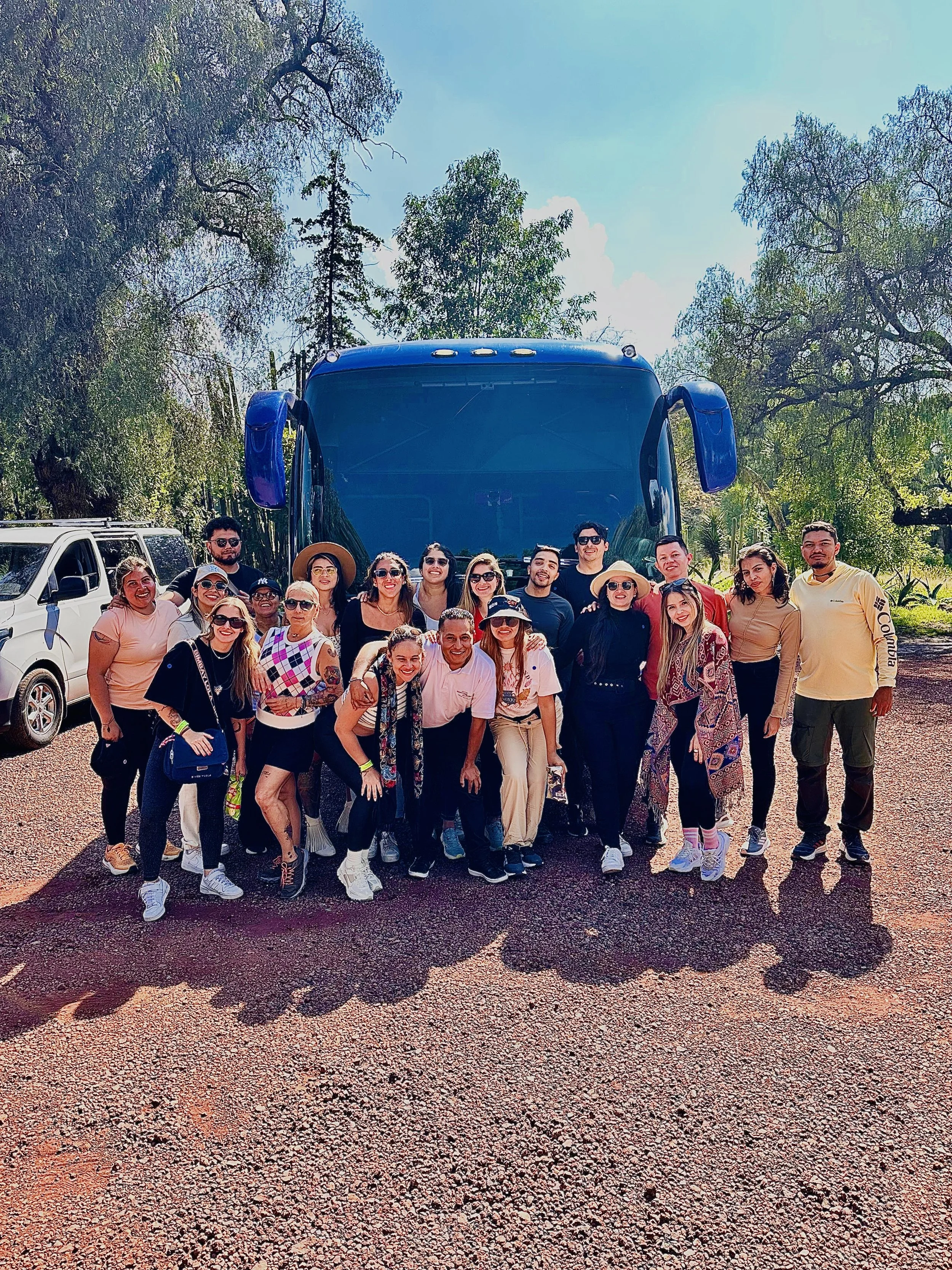 Grupo de personas sonrientes y posando frente a un autobús azul en un parque, en un día soleado.