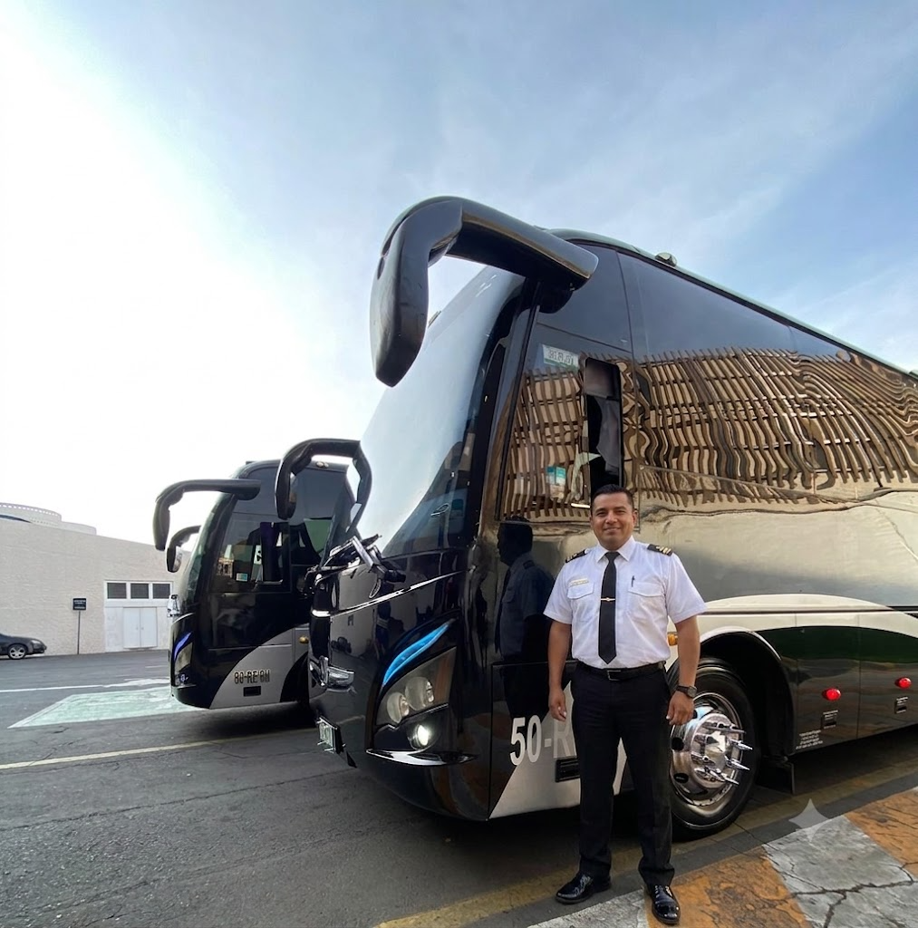 Un hombre uniformado de piloto o conductor posando frente a dos autobuses grandes en un área de estacionamiento, con cielo despejado y un edificio en el fondo.