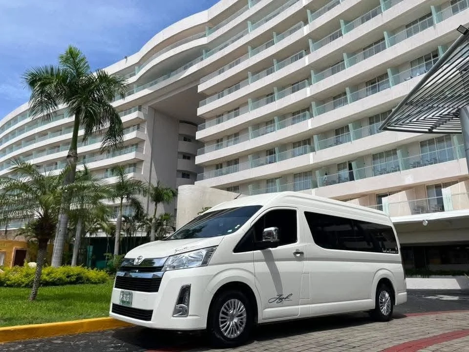 Una camioneta blanca estacionada frente a un edificio moderno de varios pisos con balcones de cristal, rodeada de palmeras y jardines.