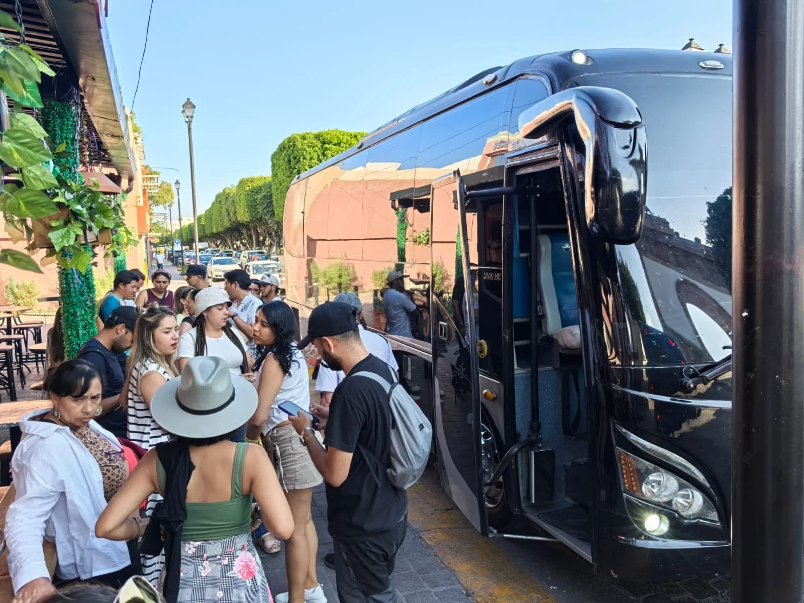 Grupo de personas haciendo fila para abordar un autobús negro en la calle, con árboles y postes de luz en el fondo.