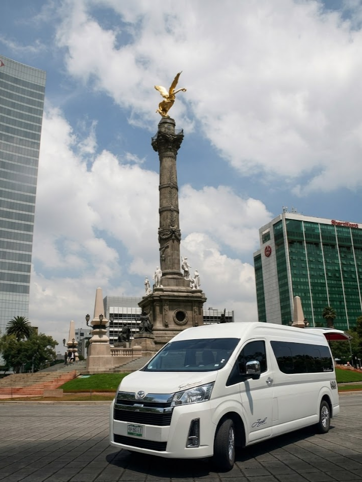 Una camioneta blanca estacionada frente a un monumento en una plaza rodeada de edificios modernos en una ciudad mexicana, con cielo parcialmente nublado.