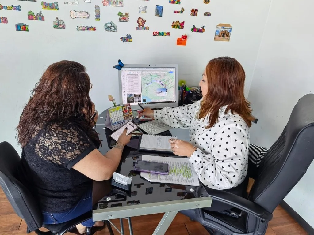 Dos mujeres en una oficina mirando una computadora, una de ellas tiene cabello rizado y la otra tiene cabello rubio, y en la pared se ven dibujos y etiquetas coloridas.