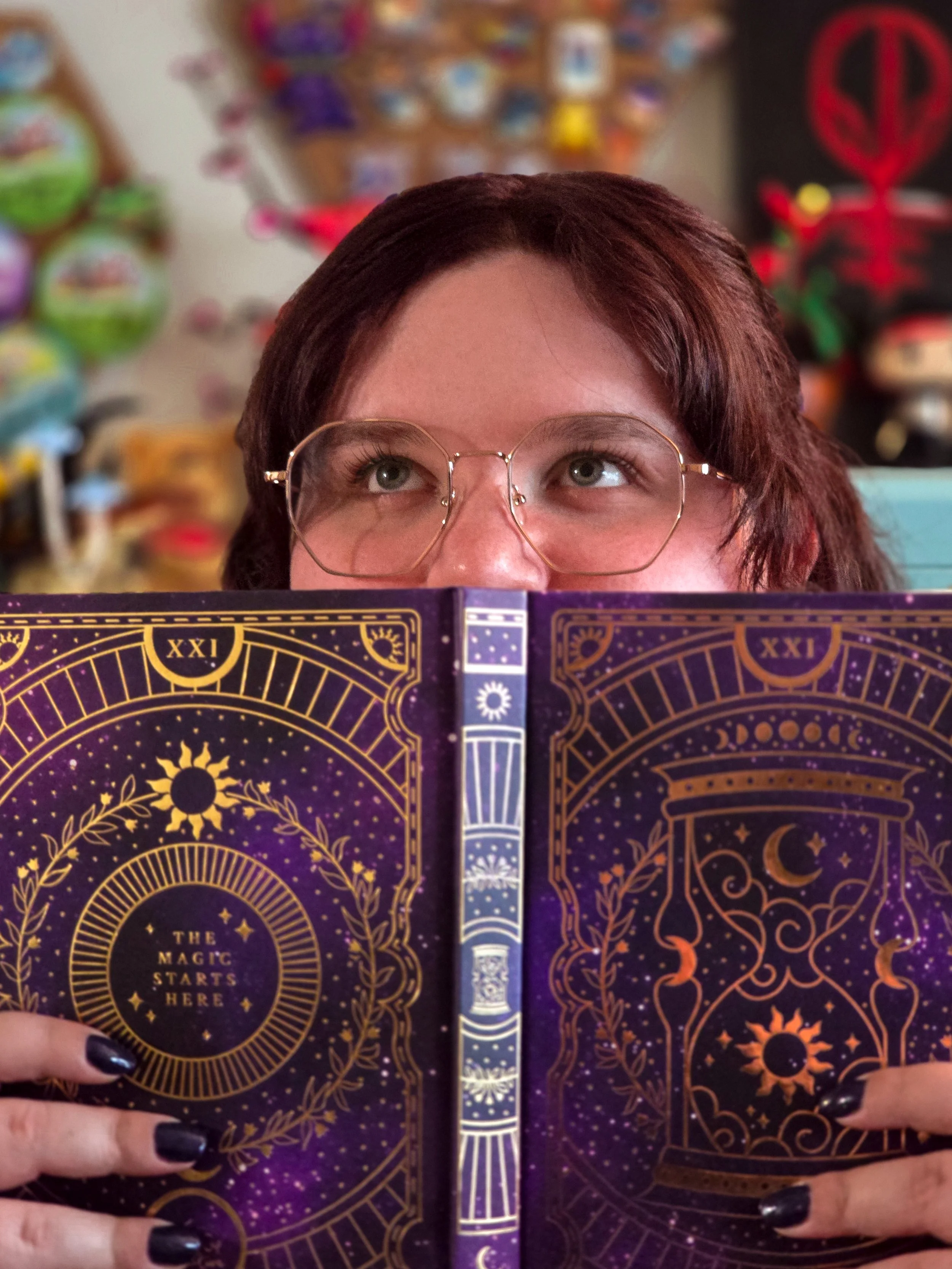 Close-up of a person with short brown hair and glasses, holding a notebook in front of their face, with a blurry, colorful background.