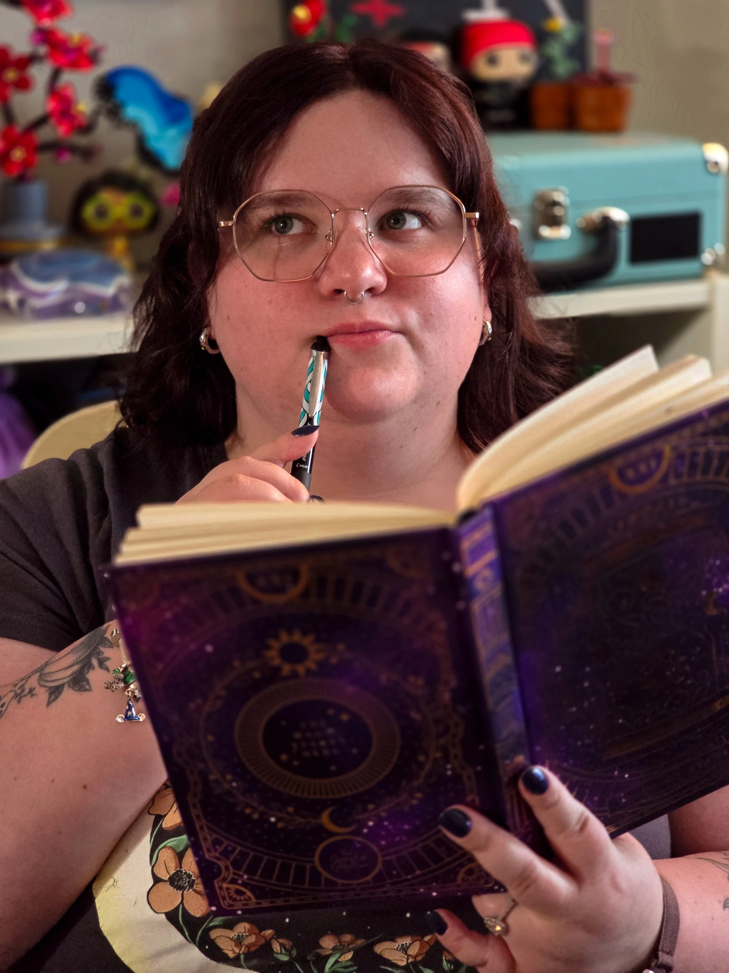A person with glasses, a septum piercing, and tattoos, holding an open notebook, with a blue pen in their mouth