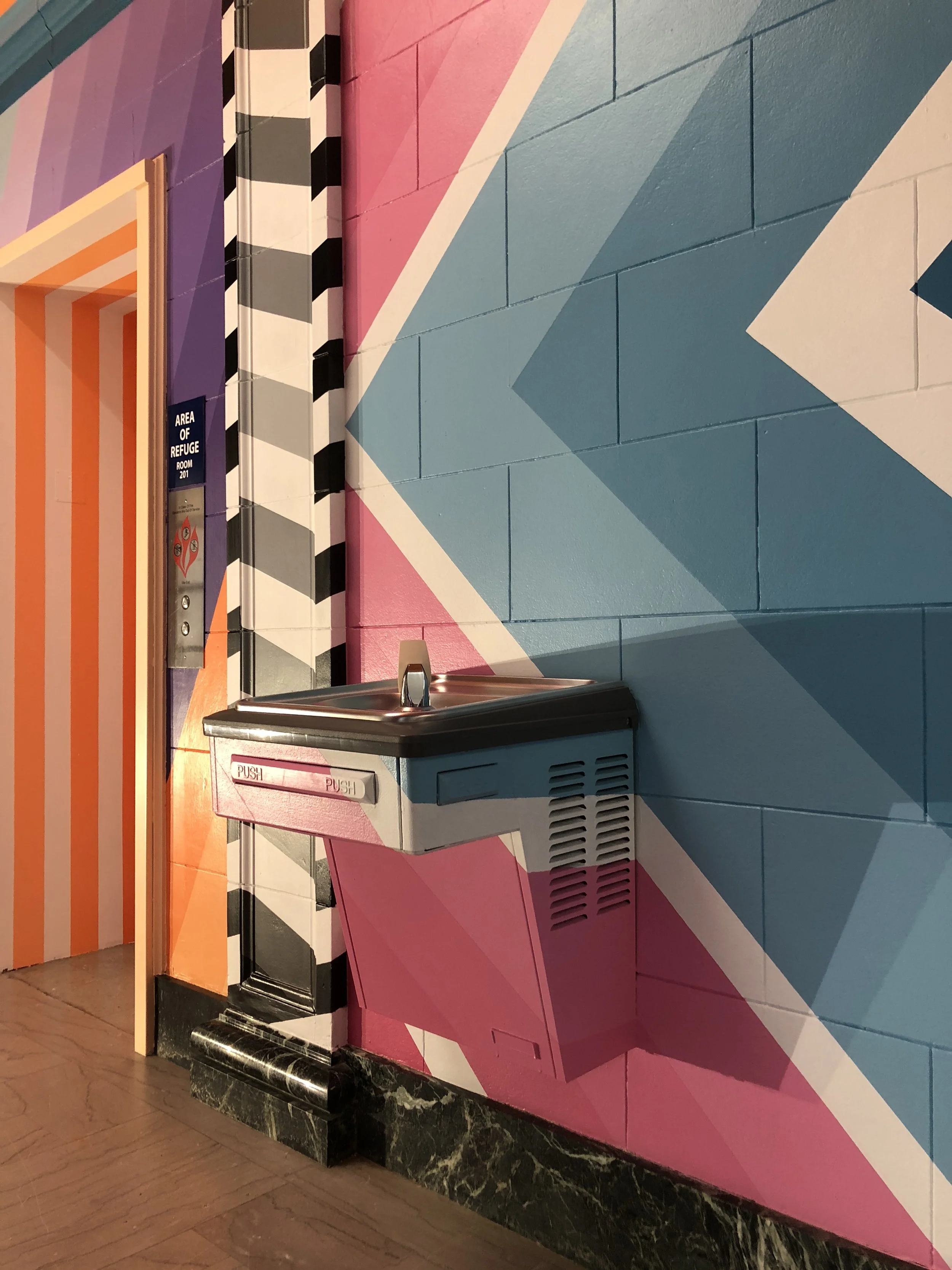Colorful geometric mural beside a drinking fountain and a door labeled “Area of Refuge Room 201.”