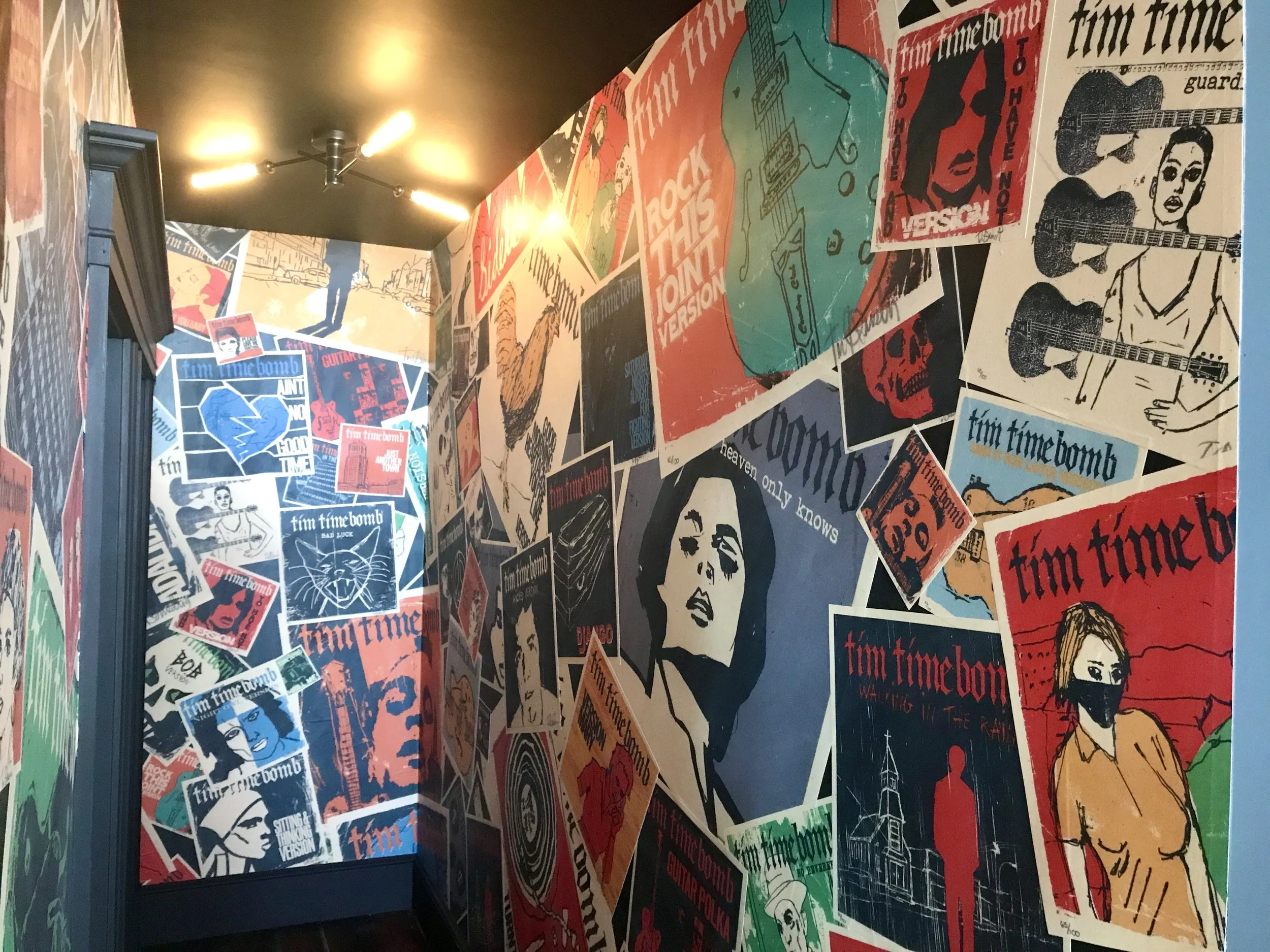 Interior hallway covered in colorful Tim Timebomb music posters and illustrated album artwork, with ceiling lights and a dark doorway.