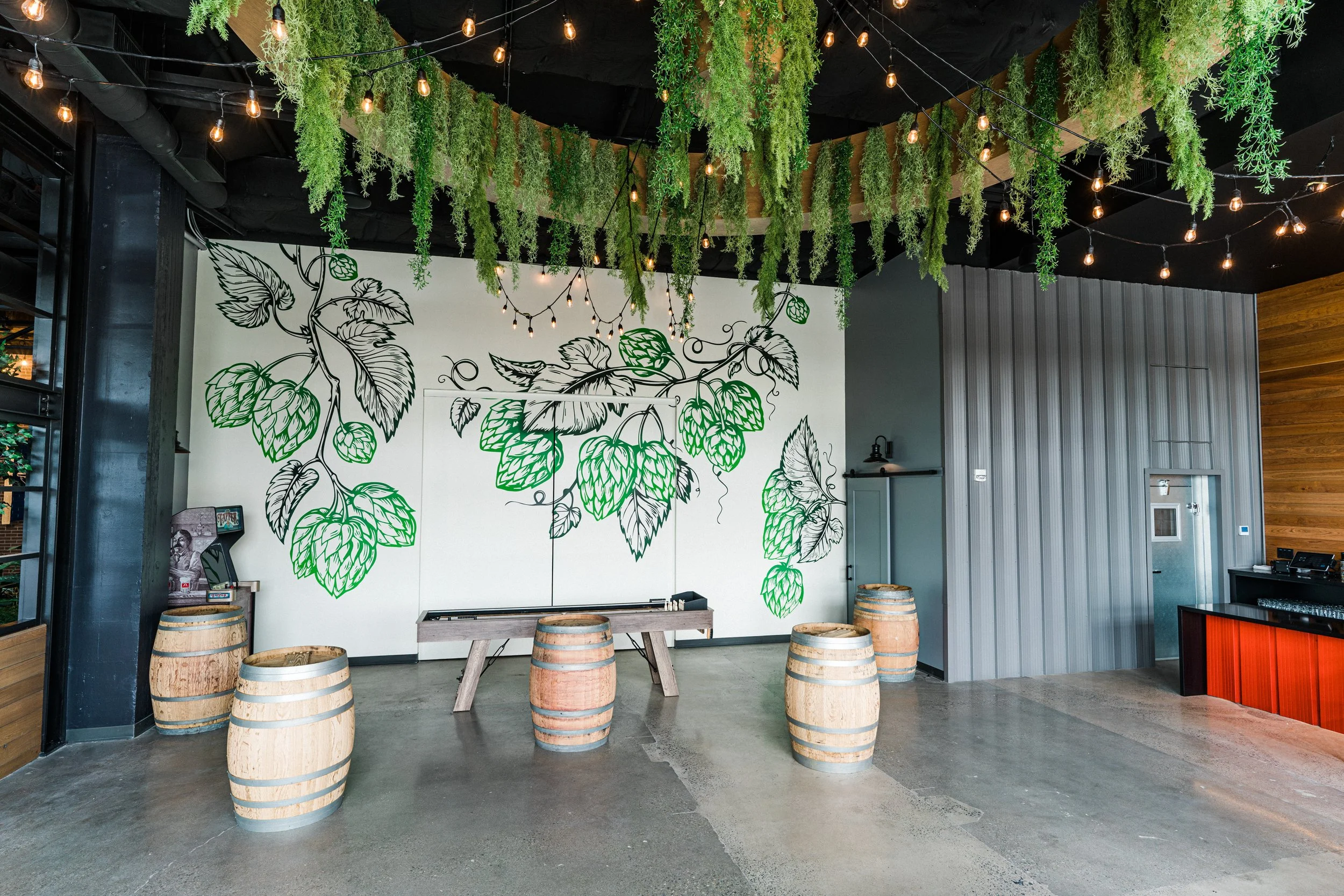 Modern brewery taproom with hop mural, hanging greenery, string lights, wooden barrels, and bar area