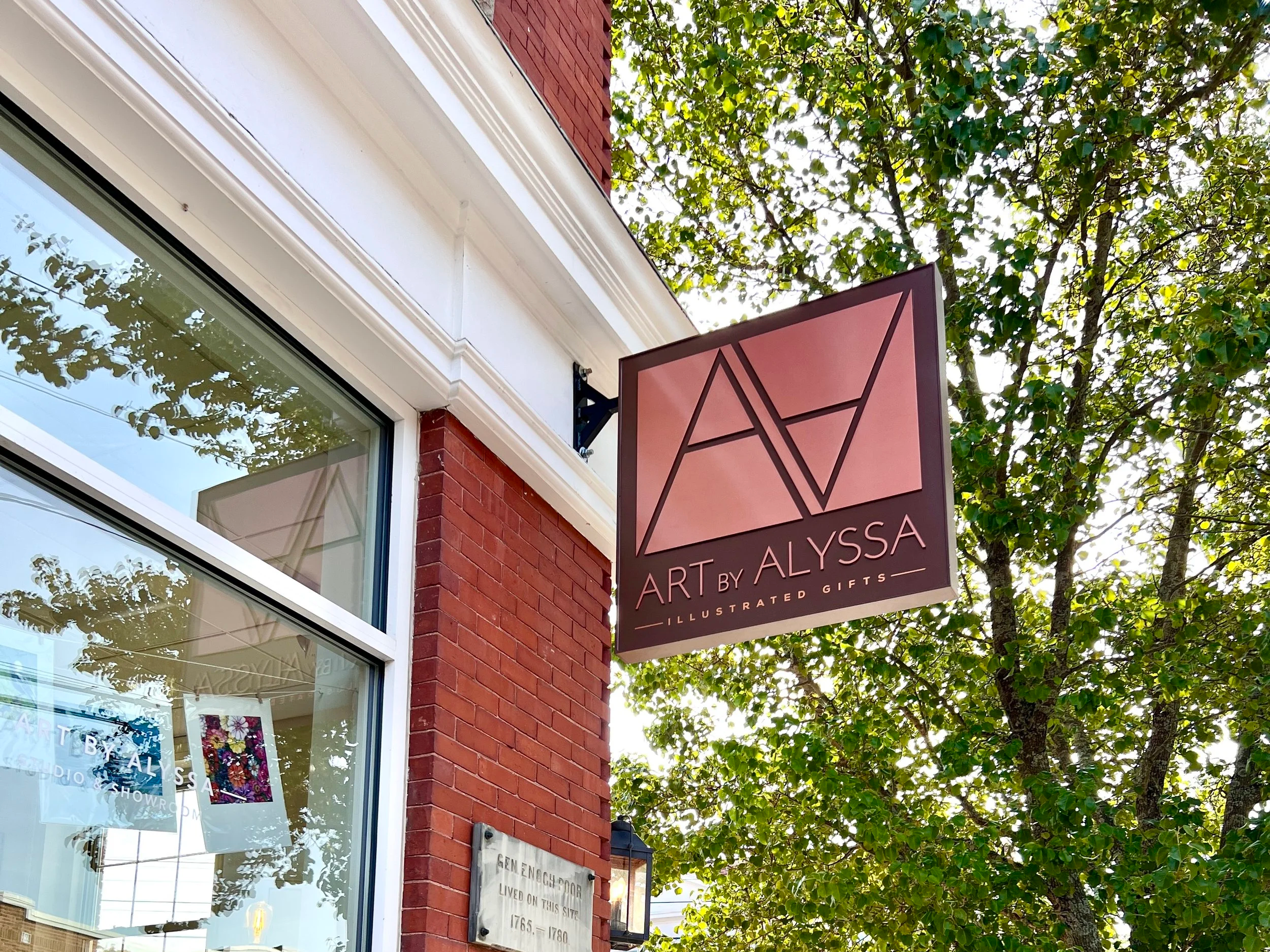 Exterior sign for Art by Alyssa illustrated gifts shop on a red brick building beneath leafy trees.