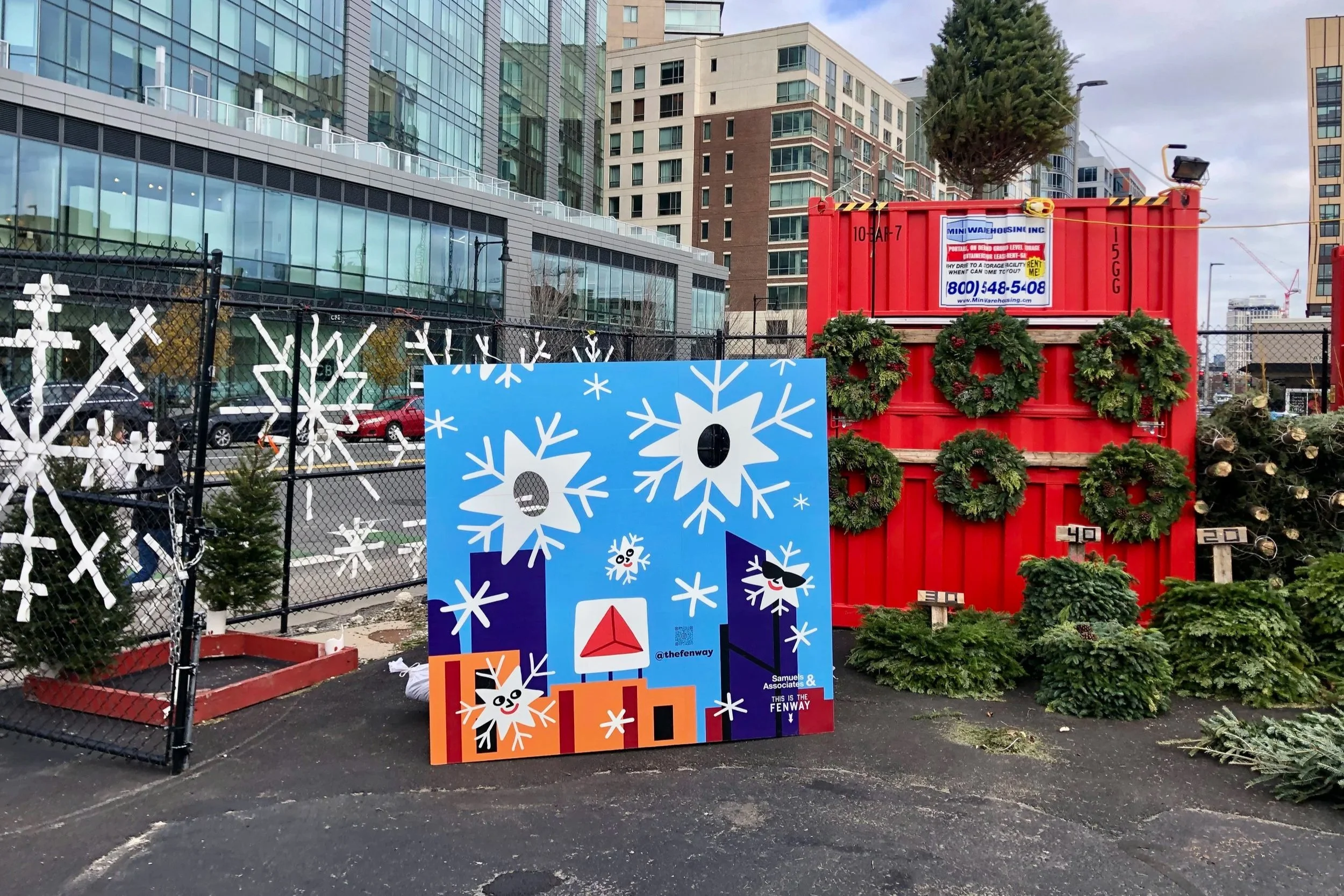 Holiday display with snowflake artwork, wreath-covered red storage container, Christmas trees, and modern city buildings
