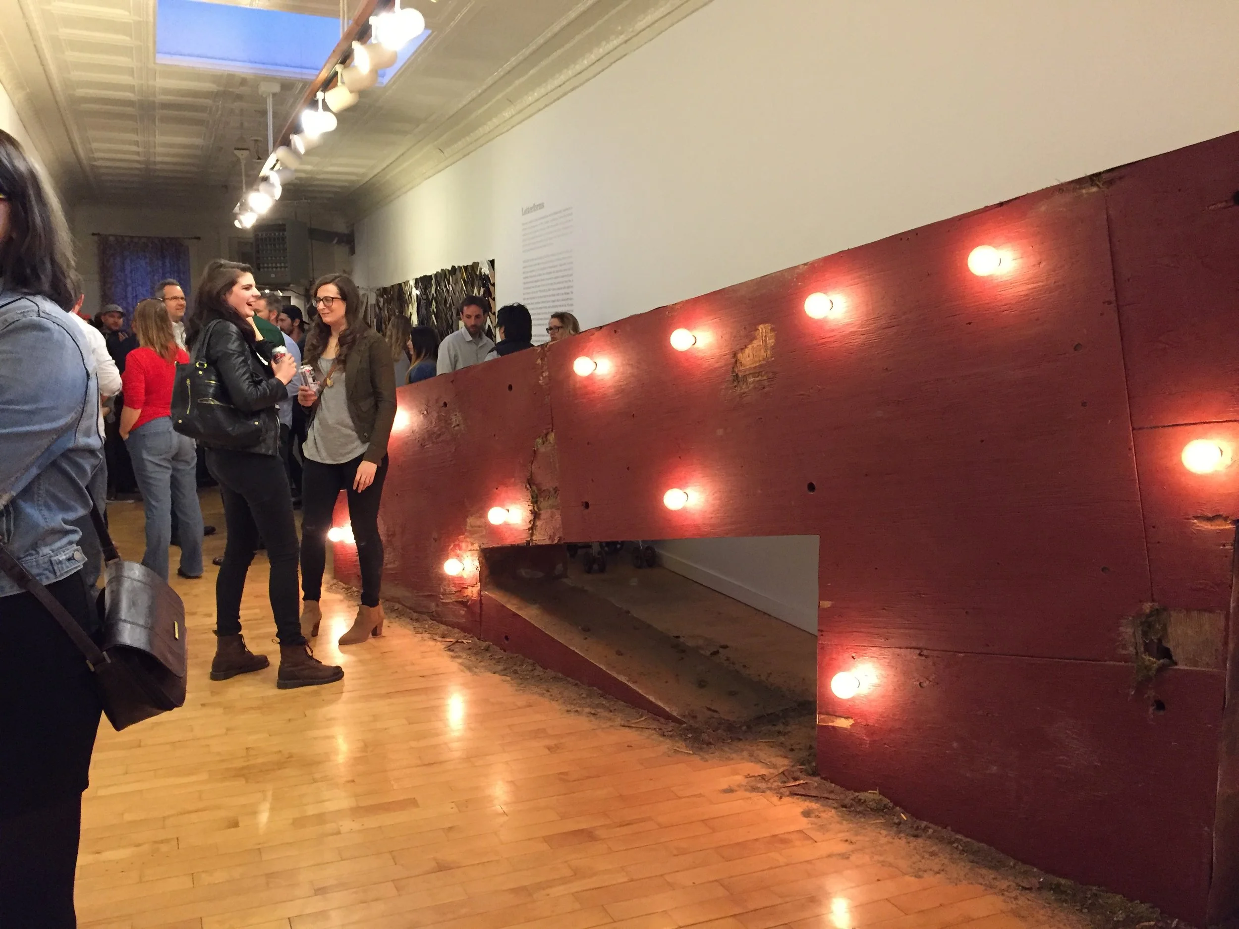 Crowded art gallery with visitors viewing a large illuminated red wooden installation.