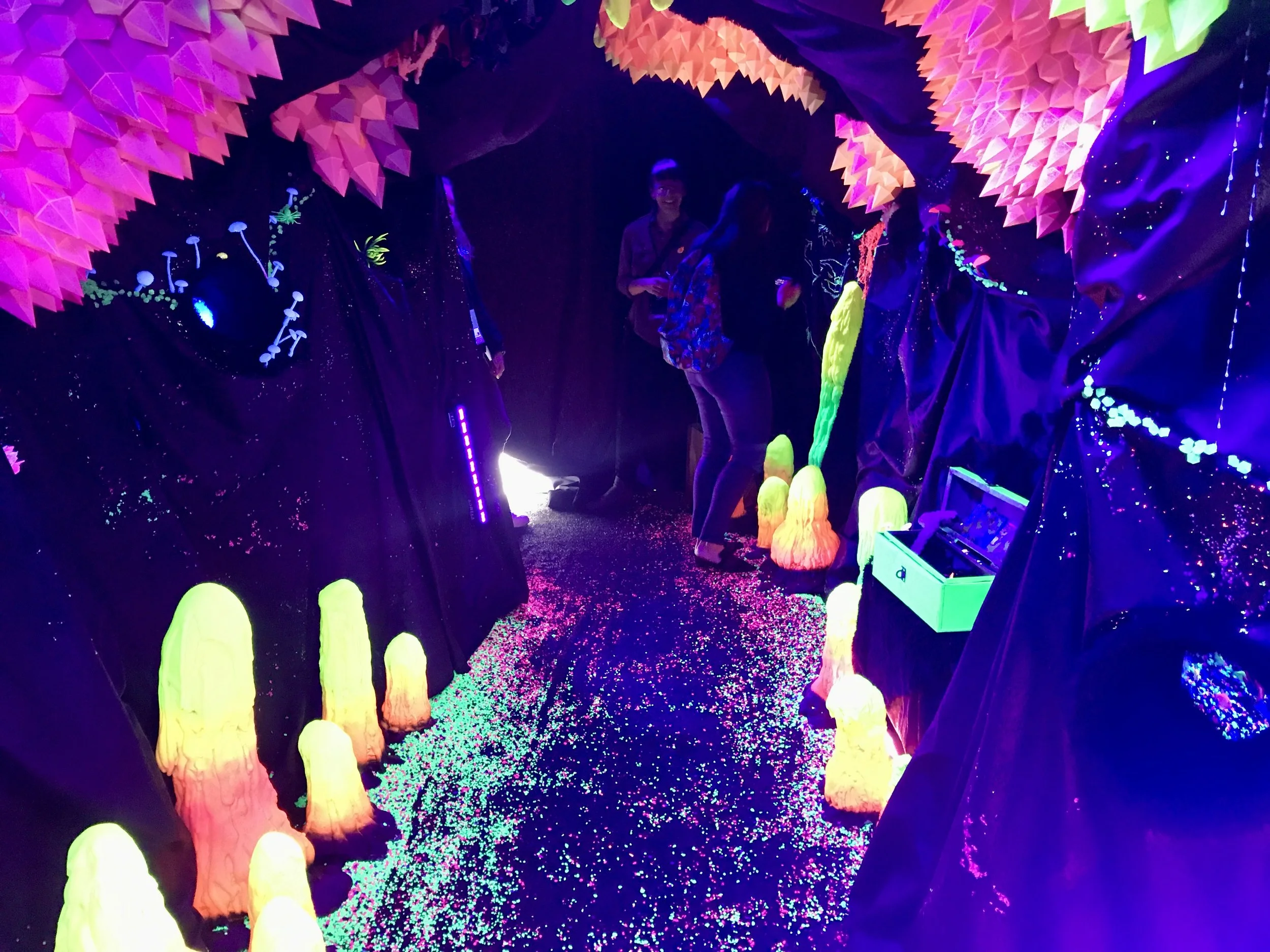 People explore a colorful blacklight installation with glowing mushrooms, crystals, and neon forest decorations.