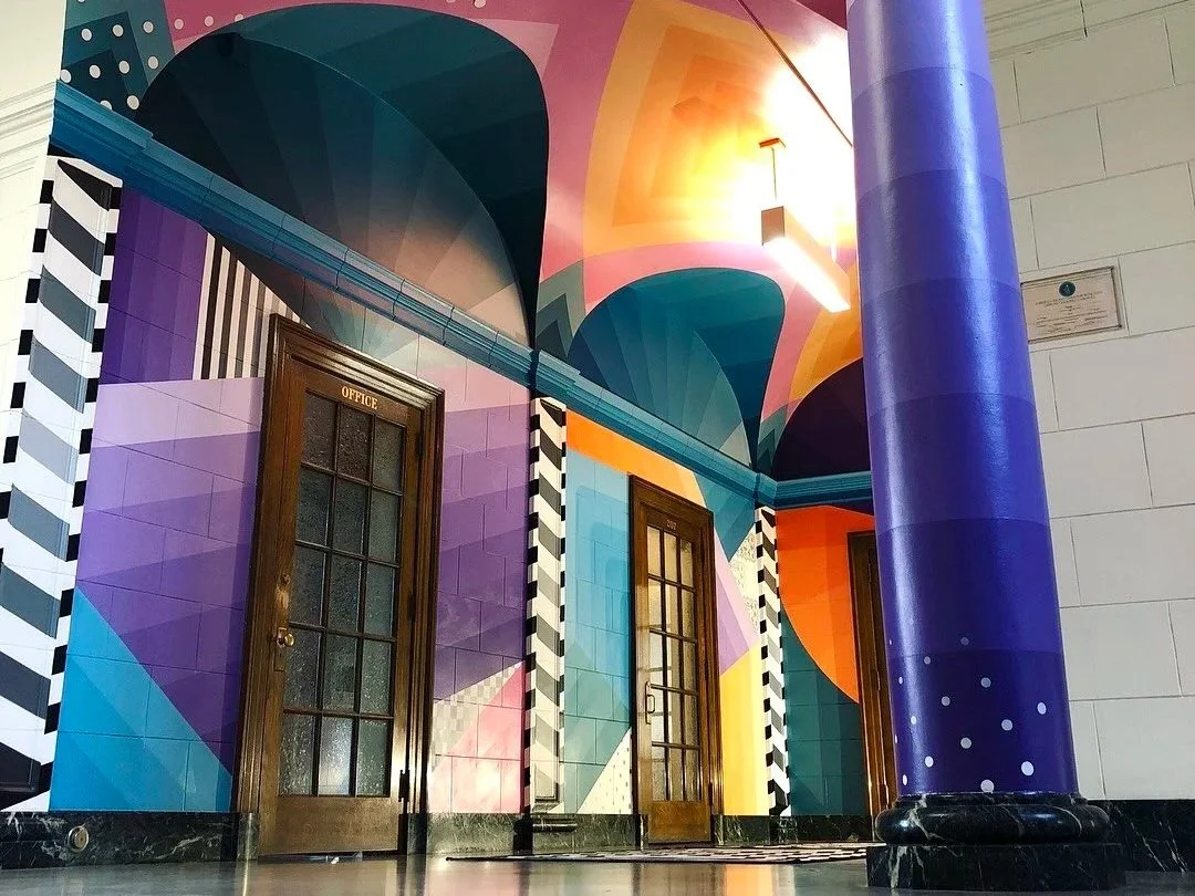 Colorful Art Deco-style interior with geometric murals, striped columns, wooden office doors, and a large purple column.