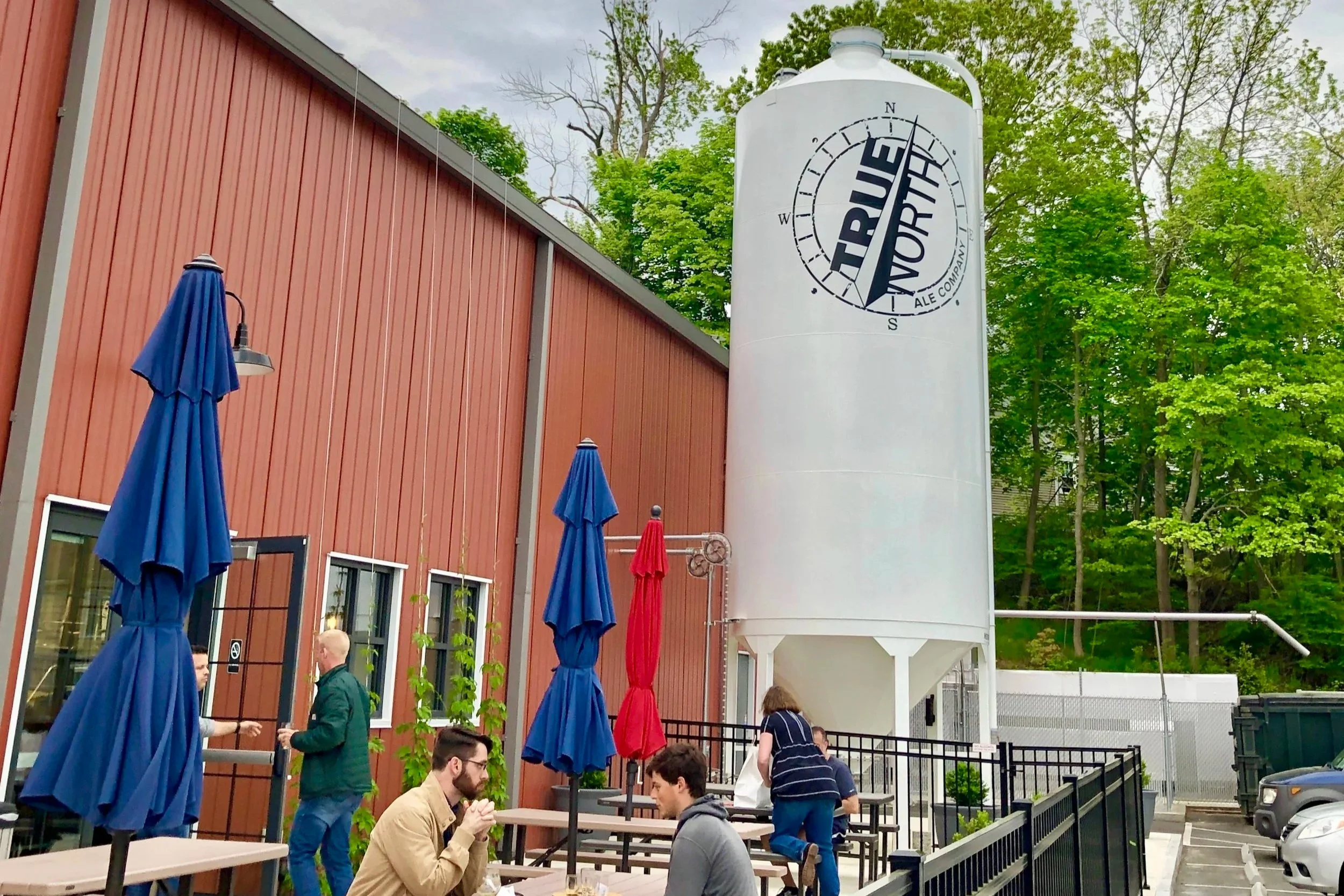 Outdoor patio at True North Ale Company featuring a large white brewery tank, red building, colorful umbrellas, picnic tables, and trees