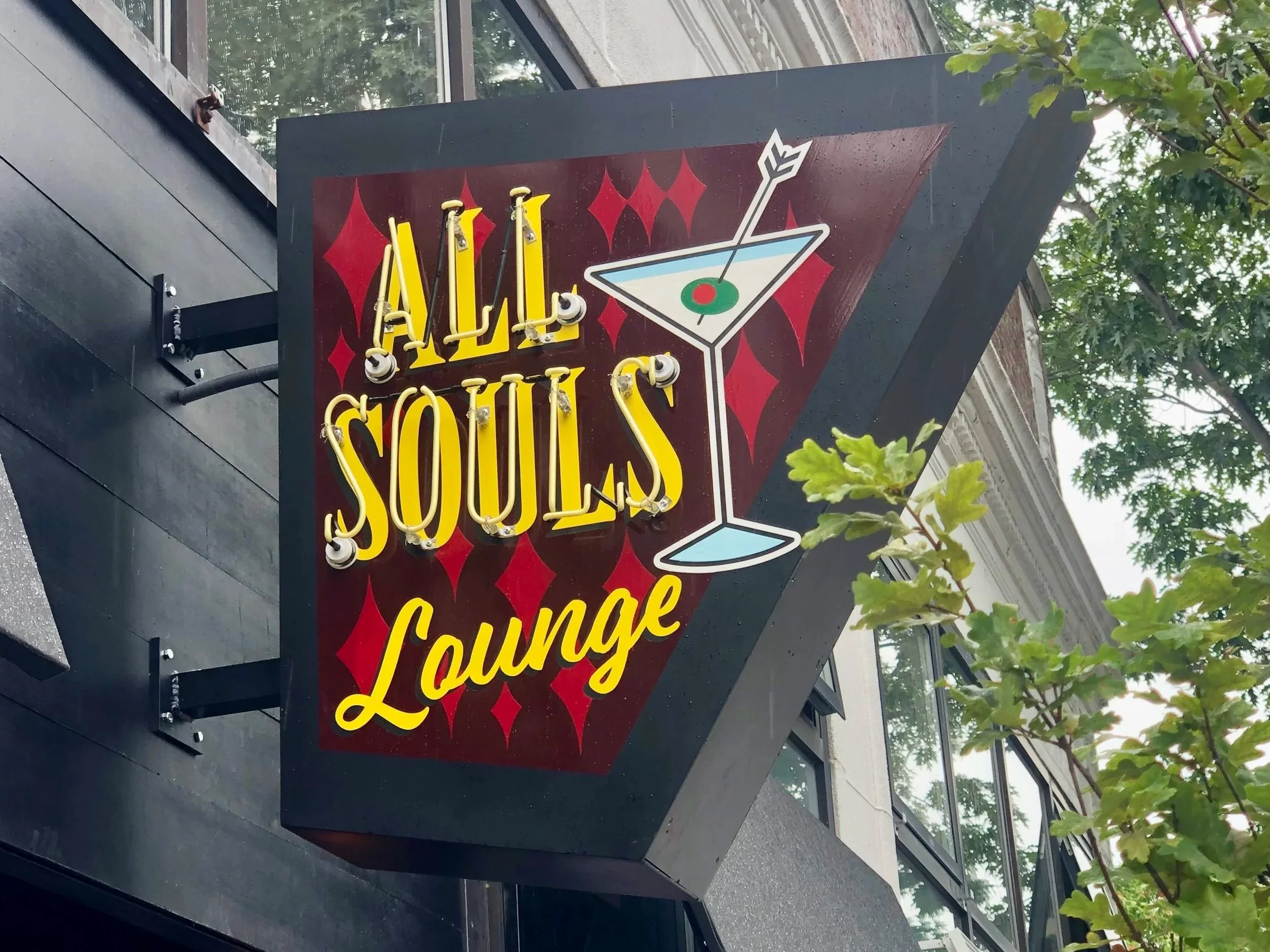 Neon sign reading “All Souls Lounge” with a martini glass and arrow outside a building.