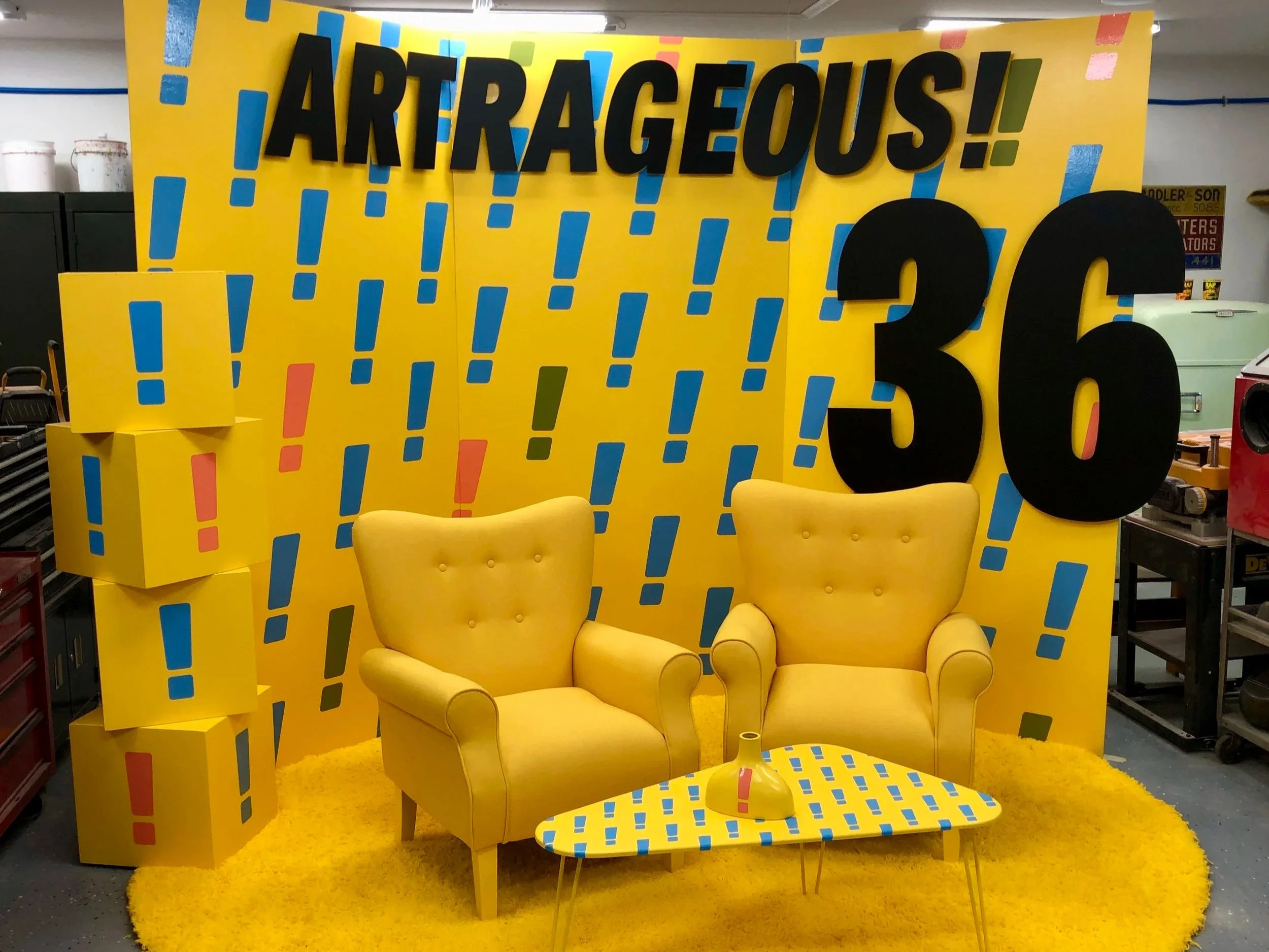 Bright yellow retro-futuristic living room display with two armchairs, patterned coffee table, geometric wall panels, and oversized black "APPLAUSE!" lettering.