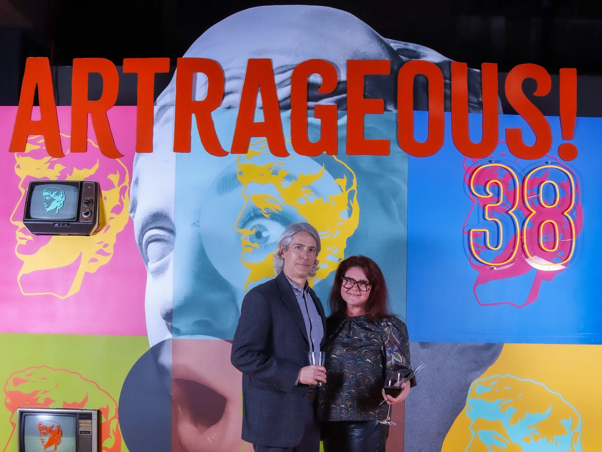 Two people pose with drinks in front of a colorful Andy Warhol-themed art display reading “ART RACECOURSE!”