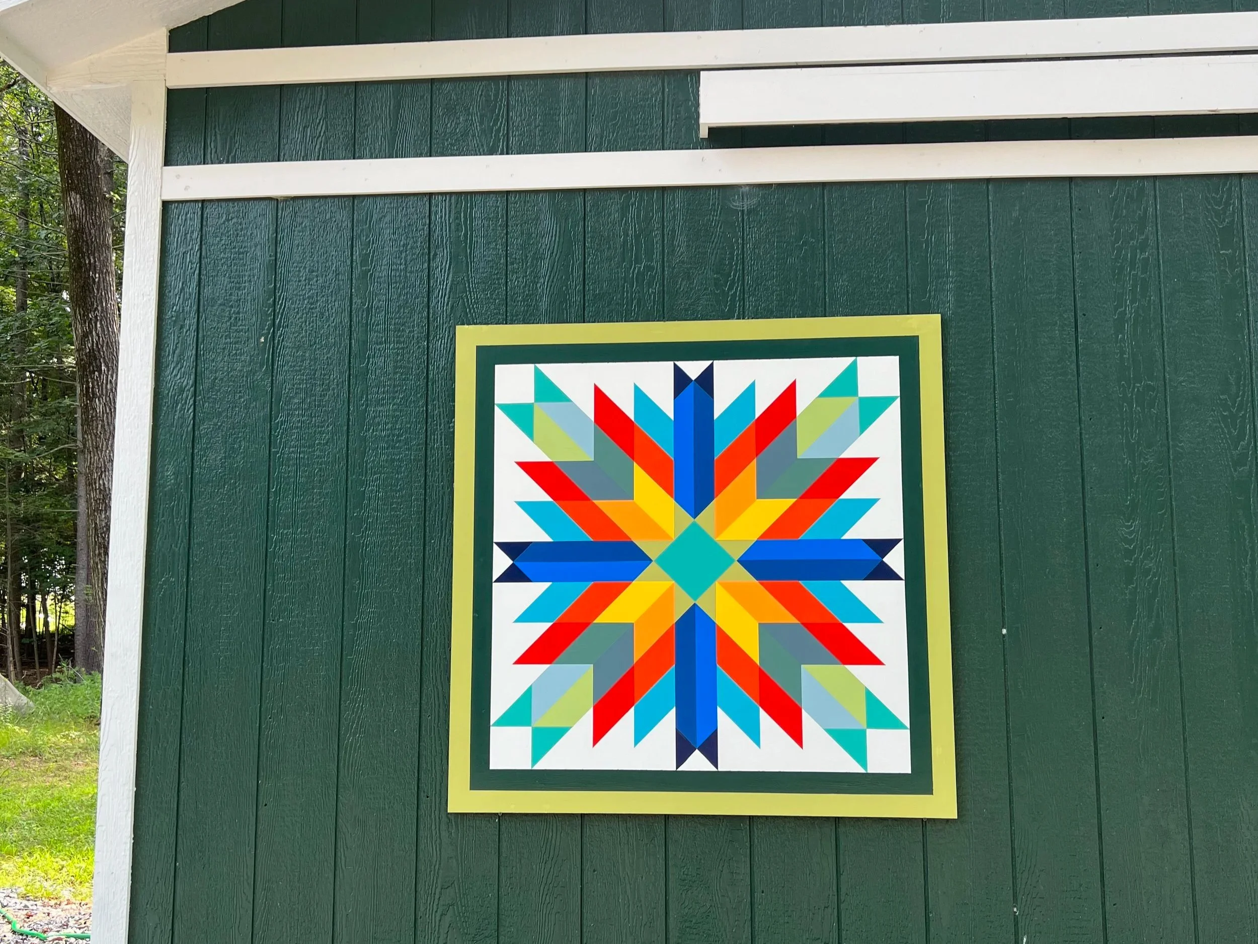 Colorful geometric quilt-block artwork mounted on a dark green shed wall outdoors