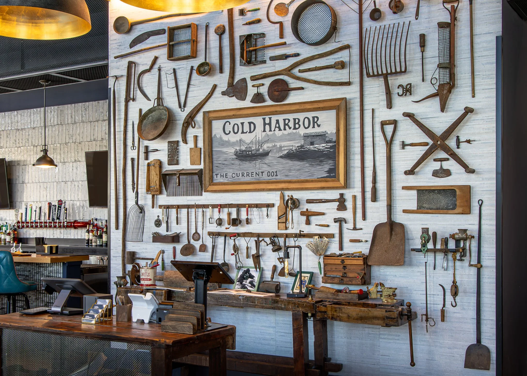 Restaurant interior with a wall display of vintage tools, farm equipment, and a framed Cold Harbor sign above the bar and workbench.