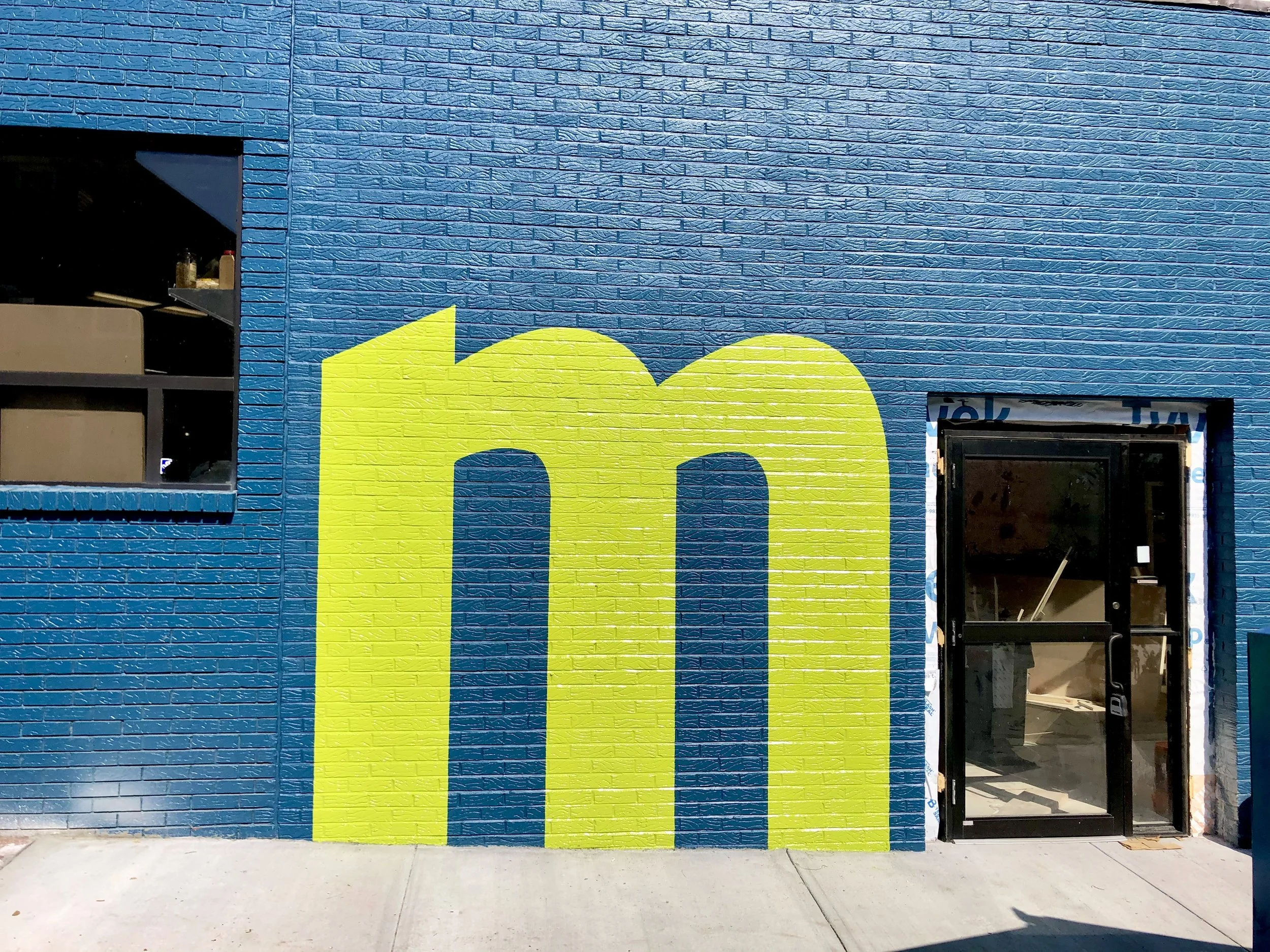 Blue brick storefront with a large yellow lowercase "m" mural and glass entrance