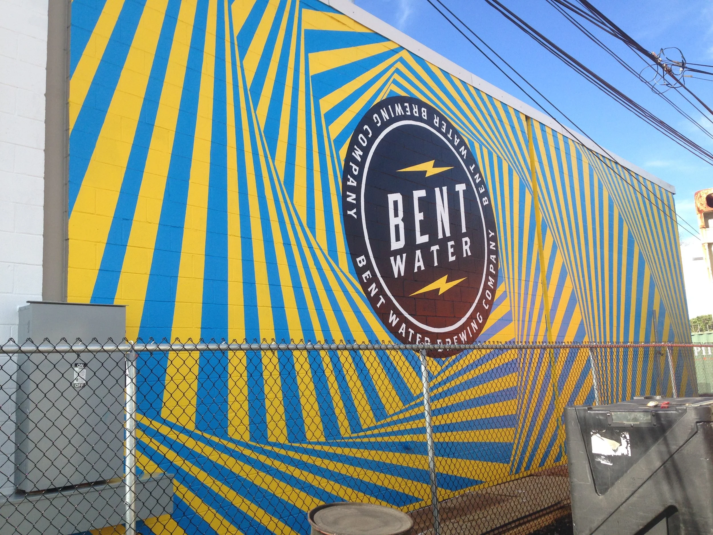 Colorful blue-and-yellow mural on a building featuring the Electric Brewing Company logo, viewed behind a chain-link fence.