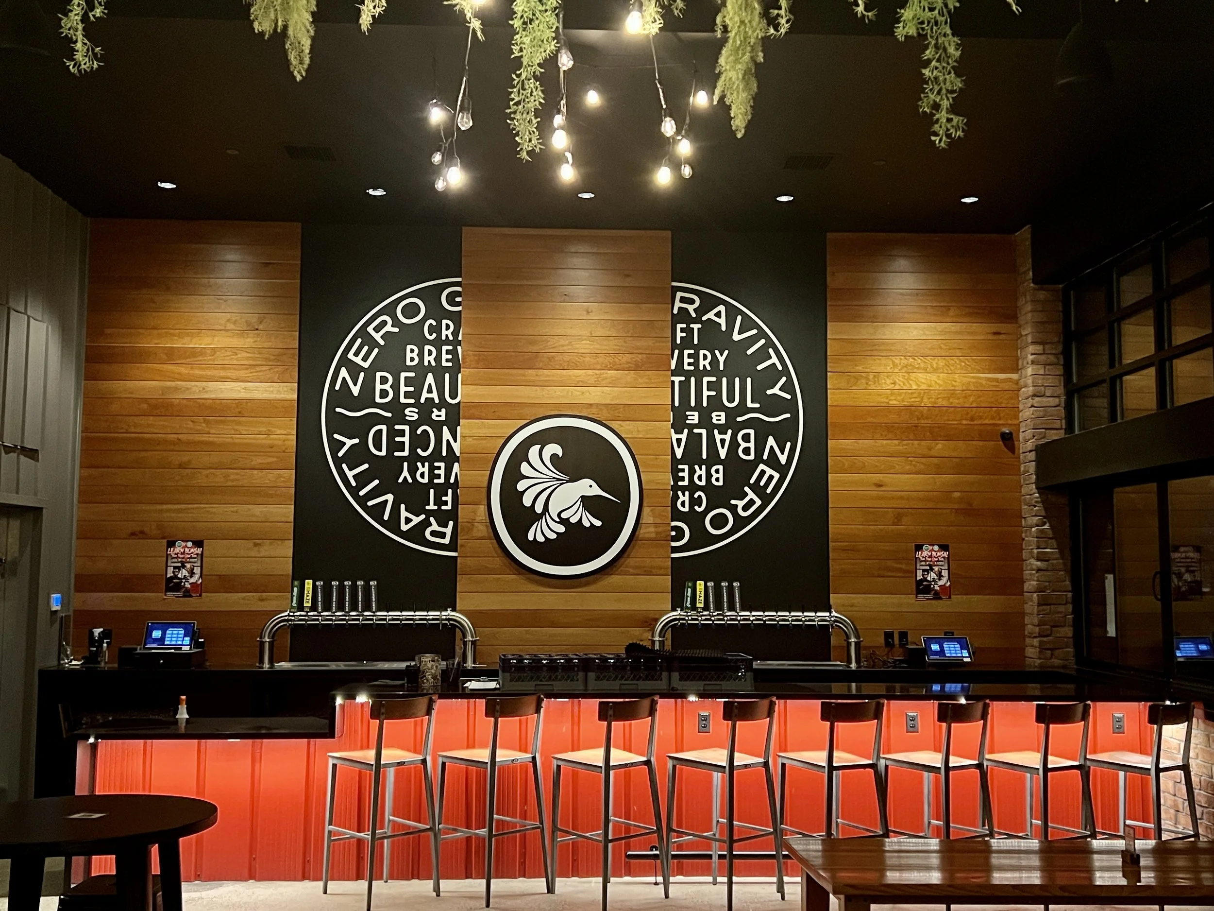 Craft brewery bar with red counter, beer taps, high-top stools, wood paneling, hanging greenery, and illuminated bird logo