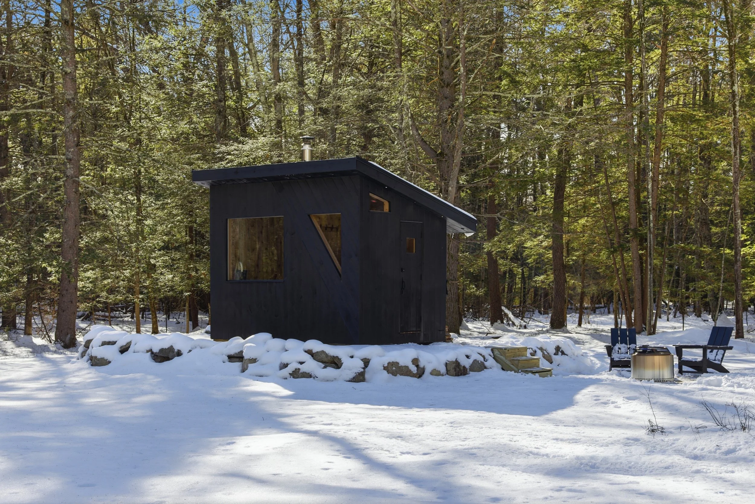 Modern black cabin with chimney, fire pit, and Adirondack chairs in a snowy forest