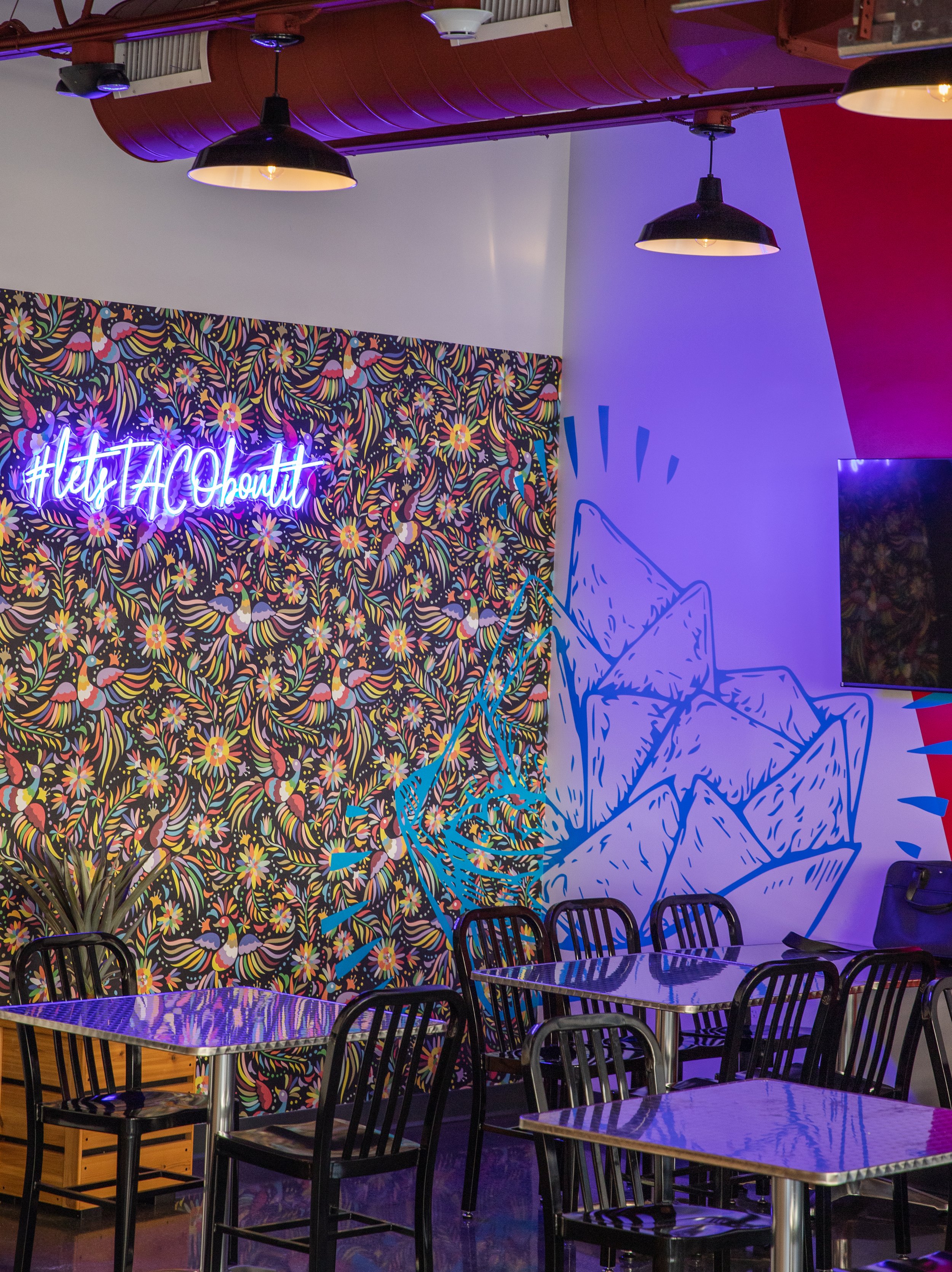 Colorful restaurant interior with floral wallpaper, purple lighting, black pendant lamps, neon “Let’s Hangout” sign, and reflective tables with chairs.