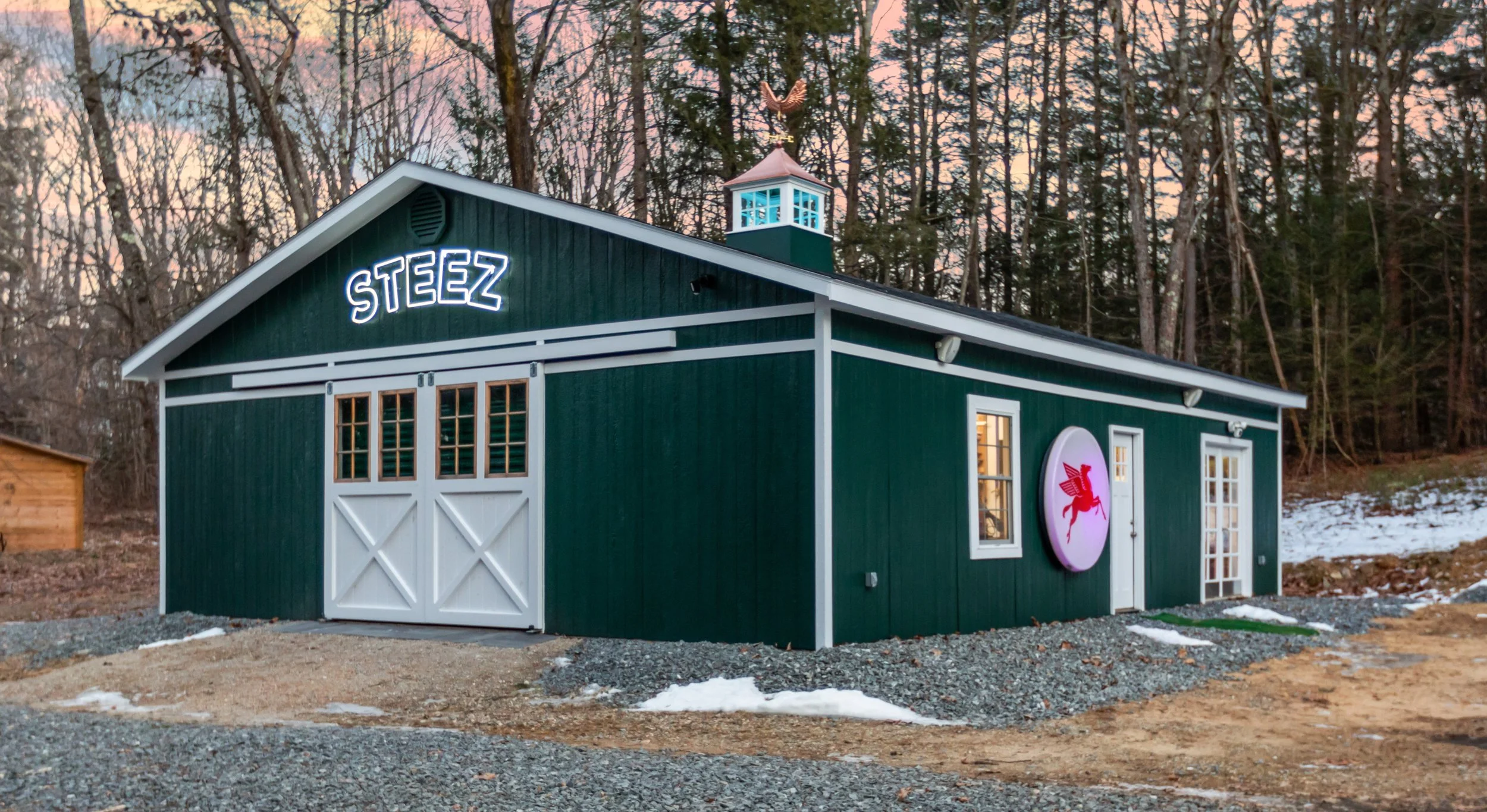 Dark green forest building labeled “STEEZ,” with white doors, cupola, and a snowboard sign in a snowy wooded setting
