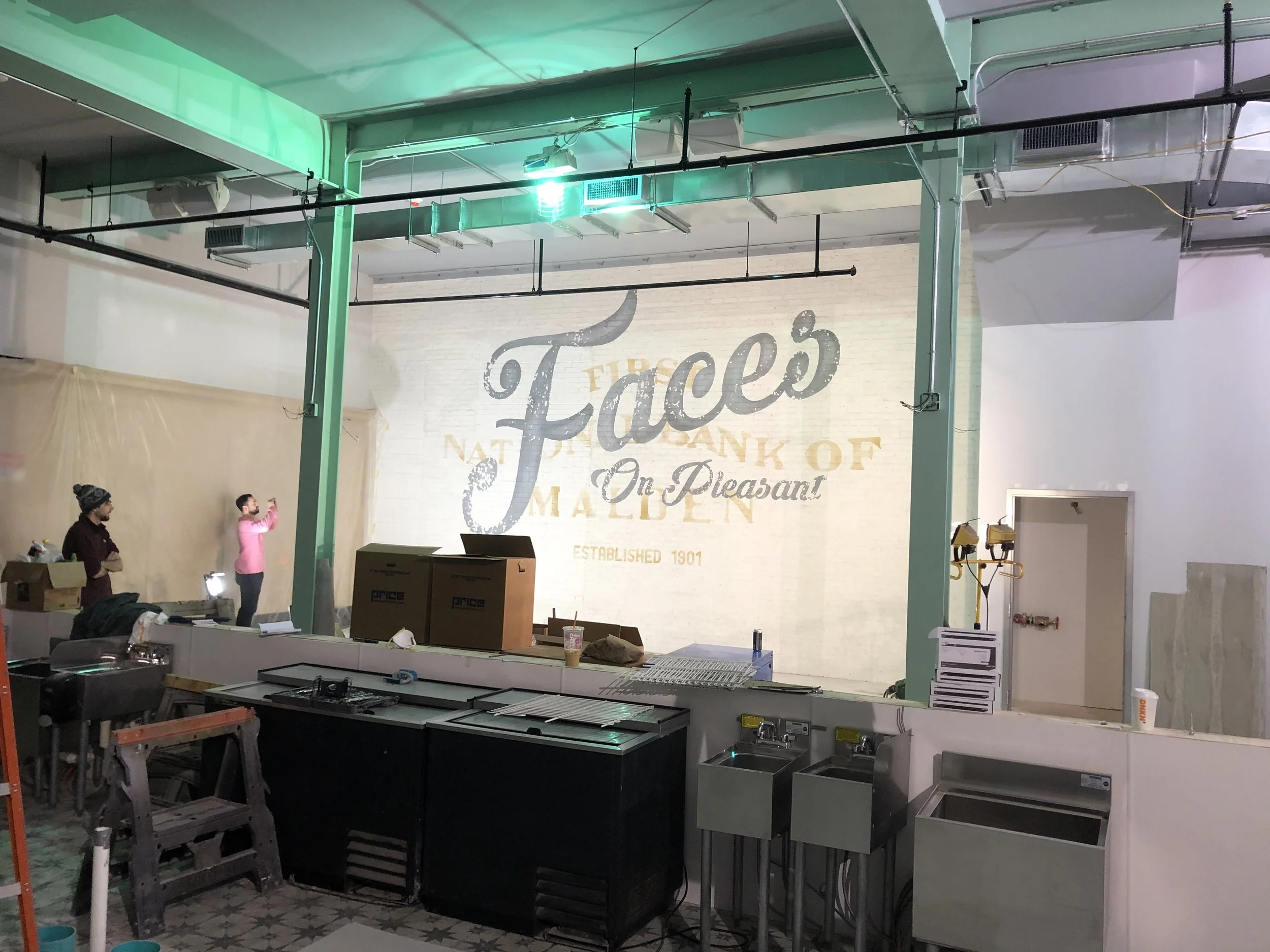 Restaurant kitchen under construction with stainless-steel equipment, workers, and a large painted Piel’s beer mural.