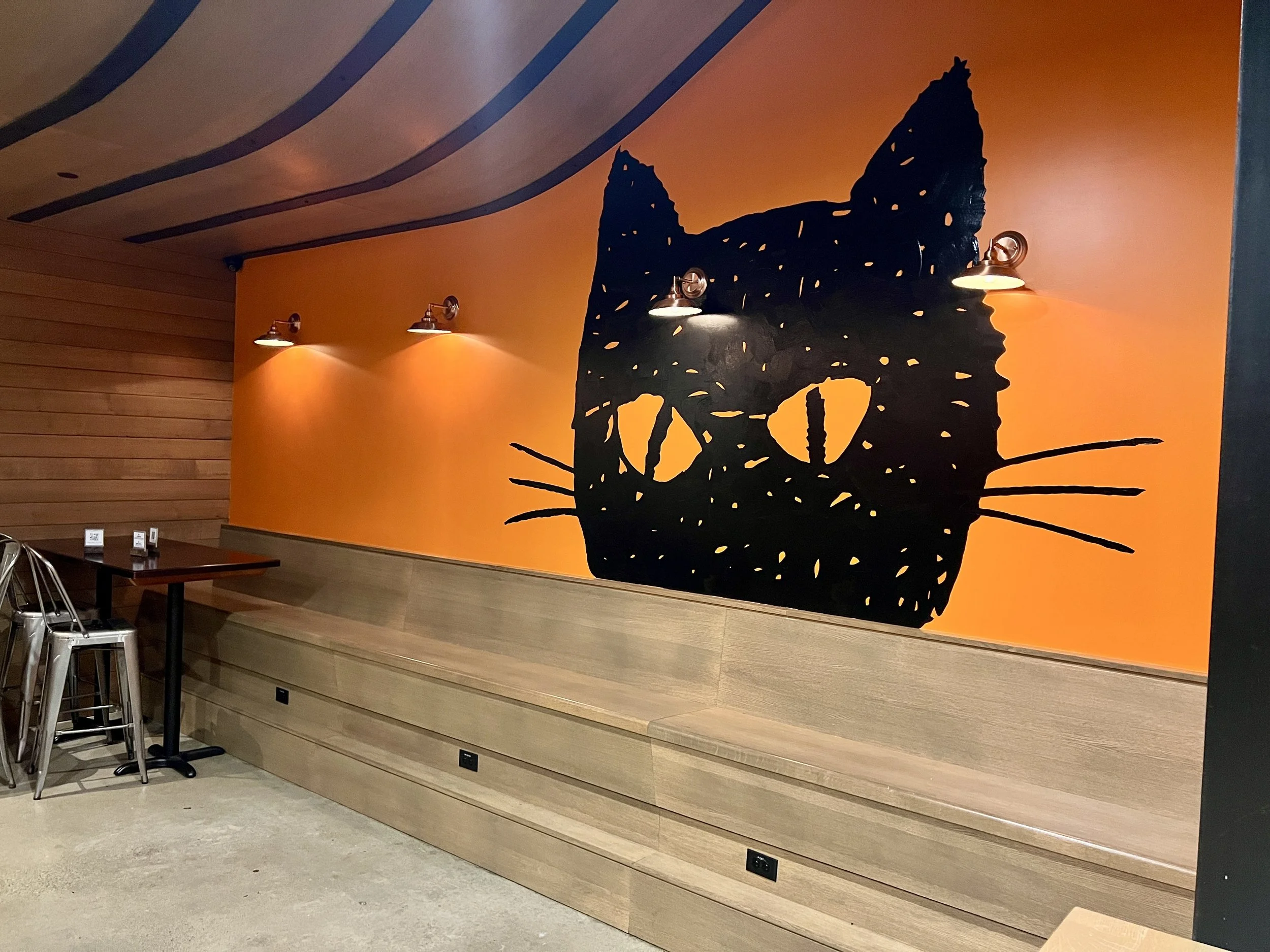 Restaurant interior with an orange wall featuring a large black cat mural, pendant lights, wood paneling, tables, and stools.