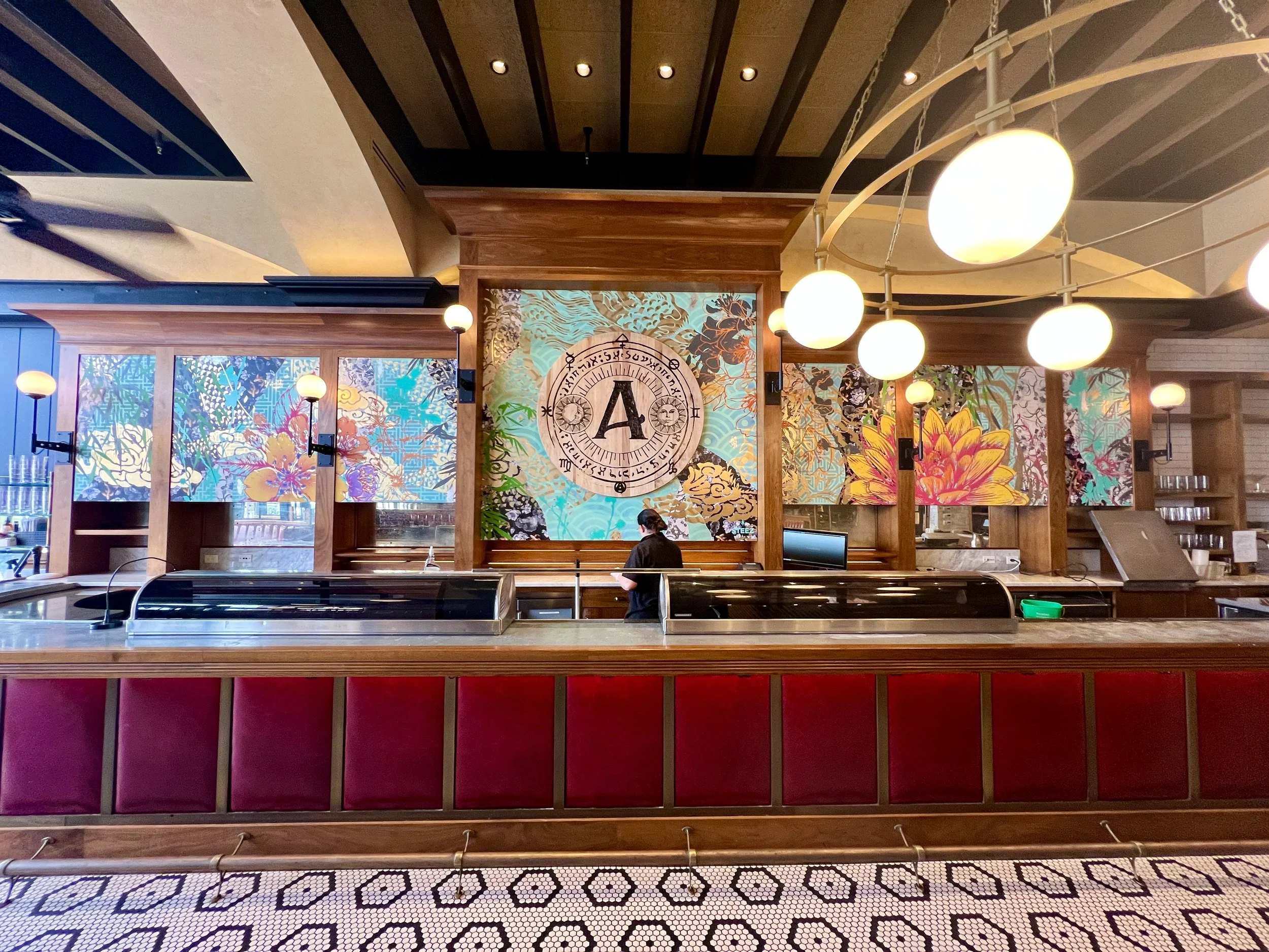 Elegant restaurant bar with red upholstered seating, colorful floral murals, globe pendant lights, and a central zodiac artwork.