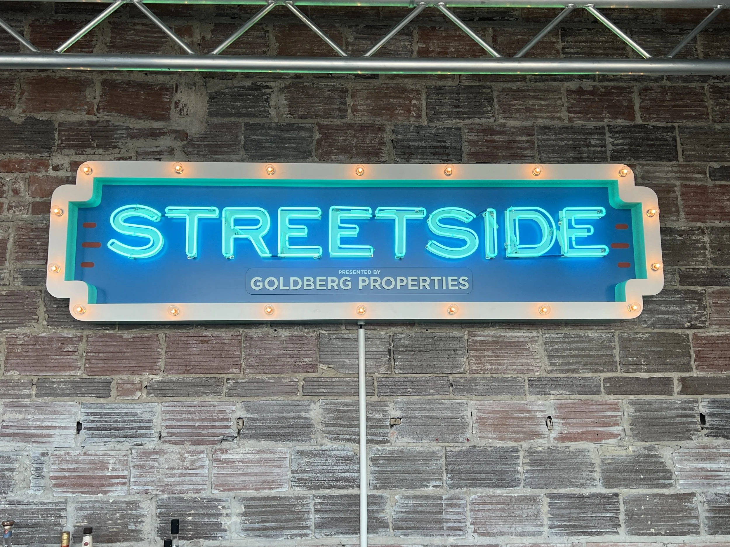 Illuminated blue neon "STREET SIDE" sign presented by Goldberg Properties on a brick wall.