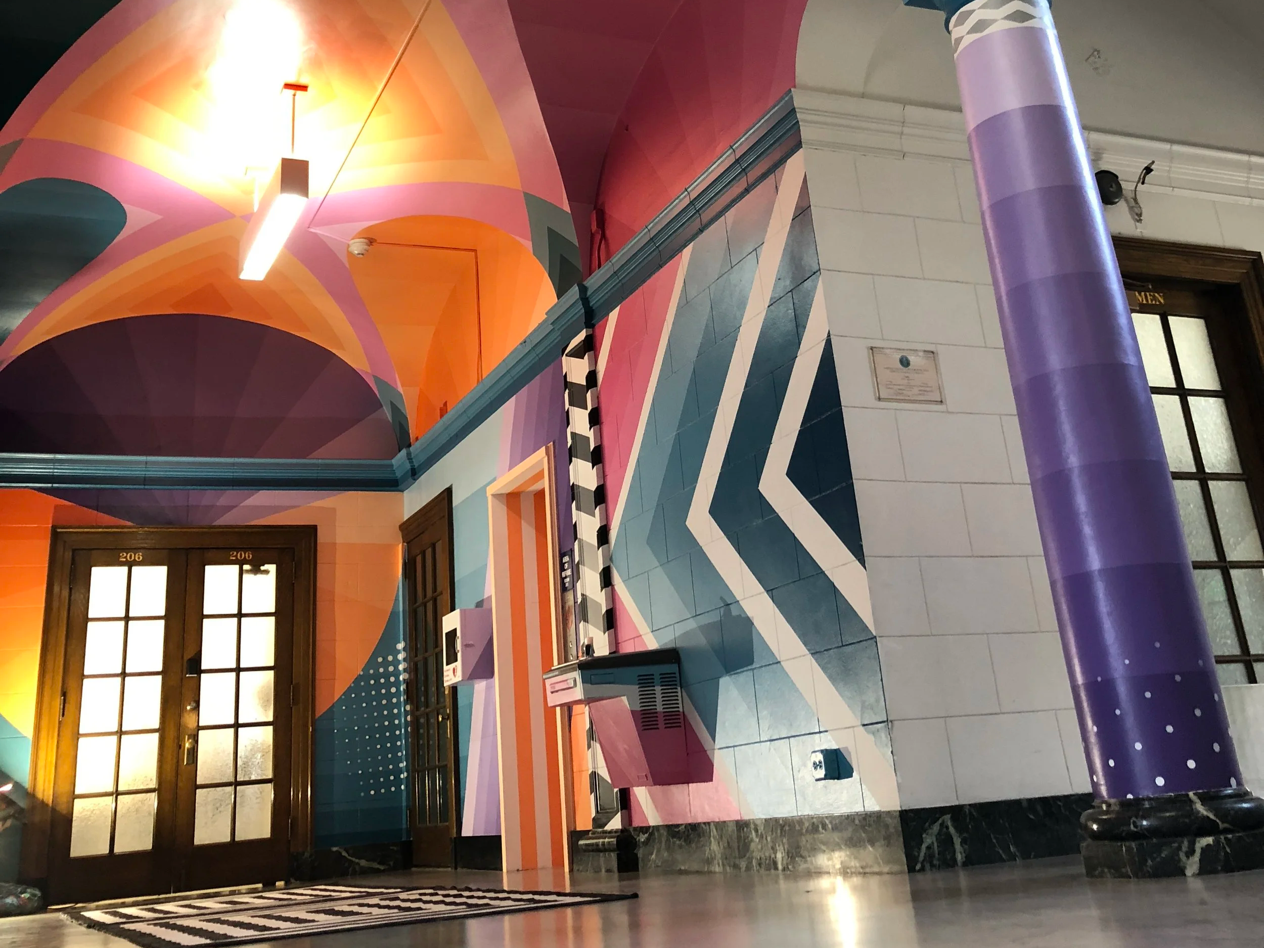 Colorful geometric mural and purple column in an ornate building hallway with numbered doors and a men's restroom entrance.