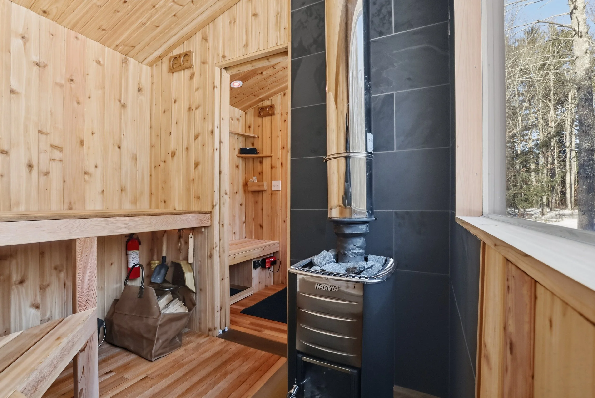 Wood-paneled sauna interior with a Harvia wood-burning stove, benches, firewood, and forest view through the window