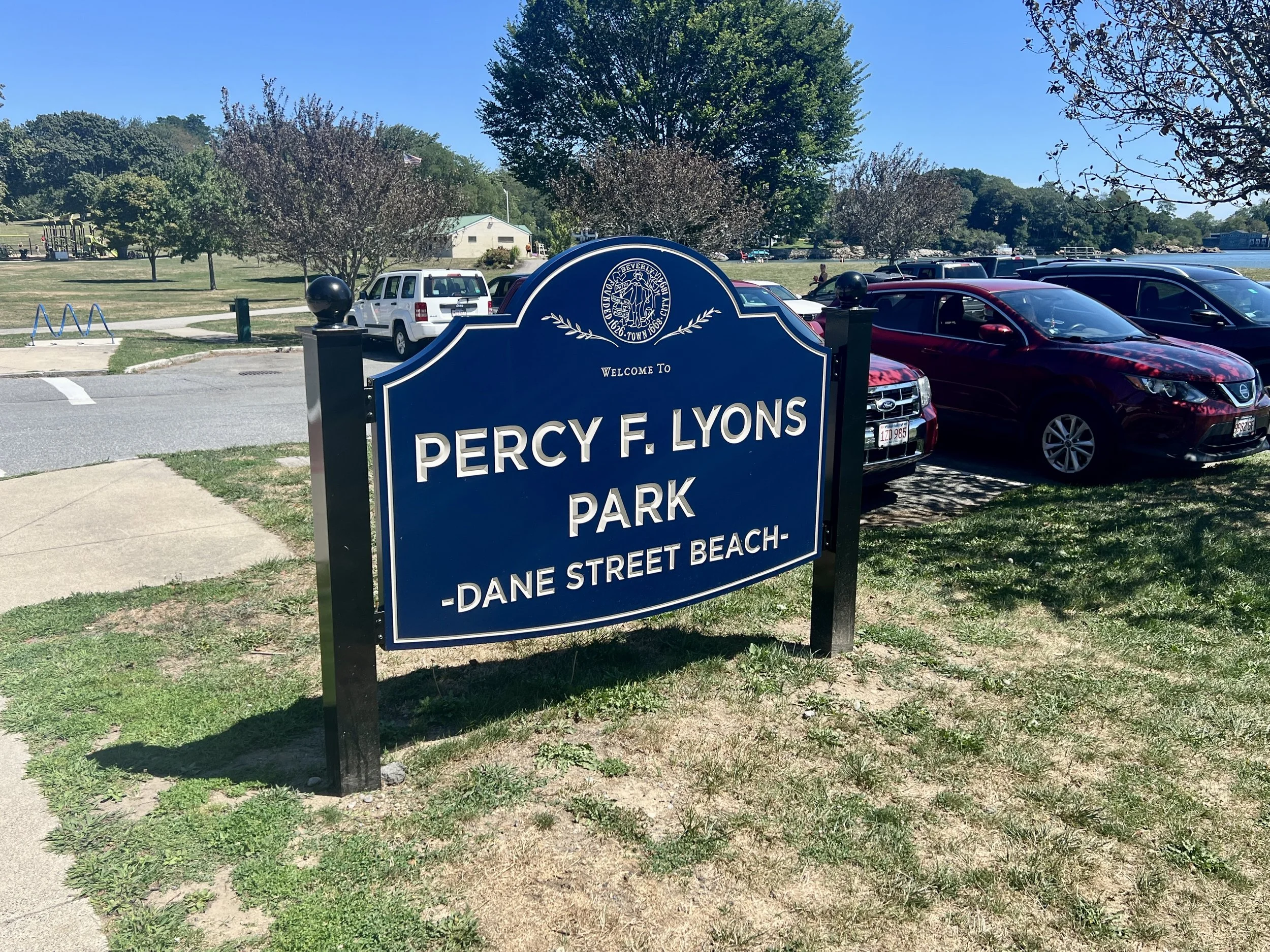 Welcome to Percy F. Lyons Park sign at a lakeside beach with trees, playground, and parked cars.