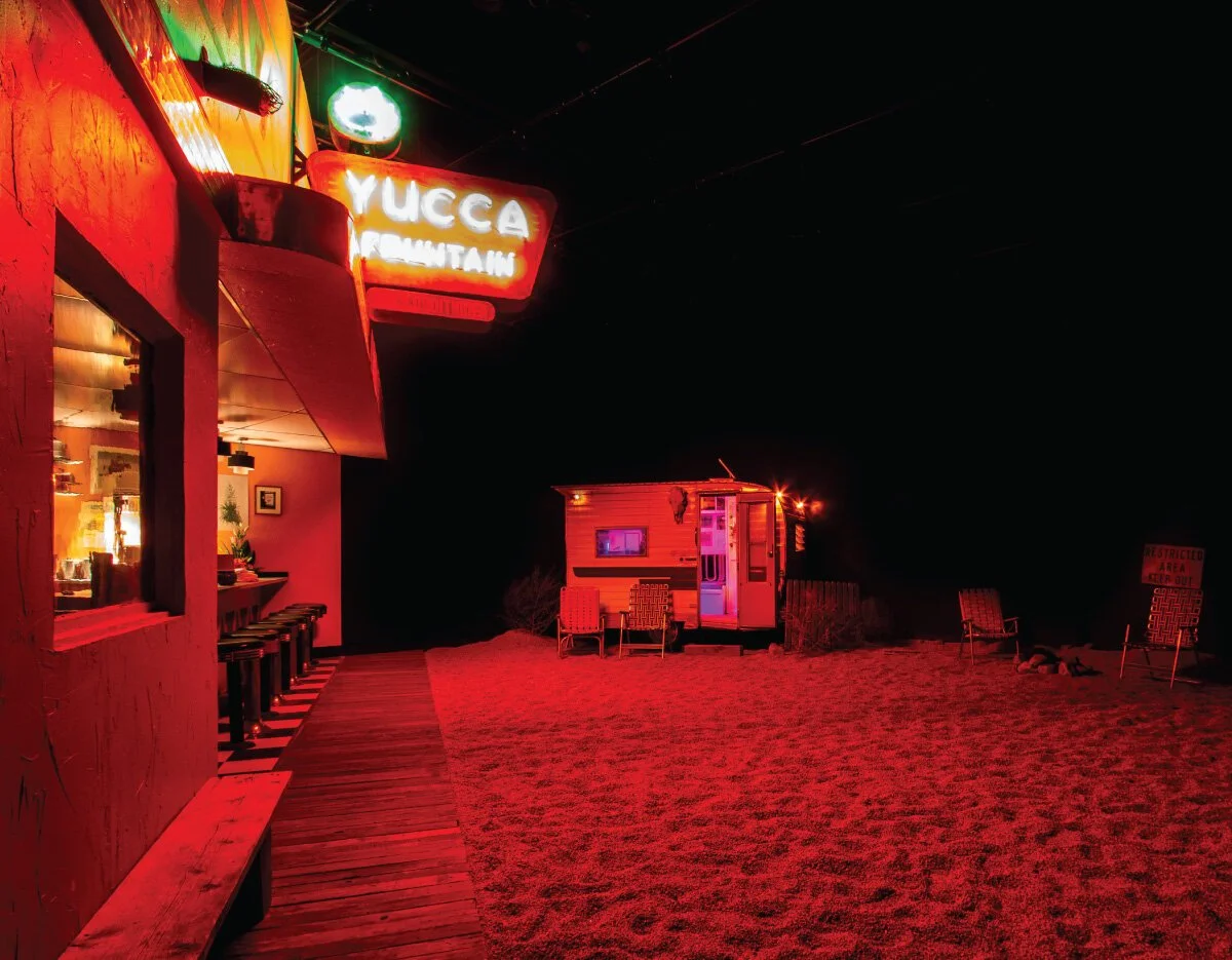 Yucca Mountain bar exterior glowing with red neon lighting at night, featuring a trailer and outdoor chairs.