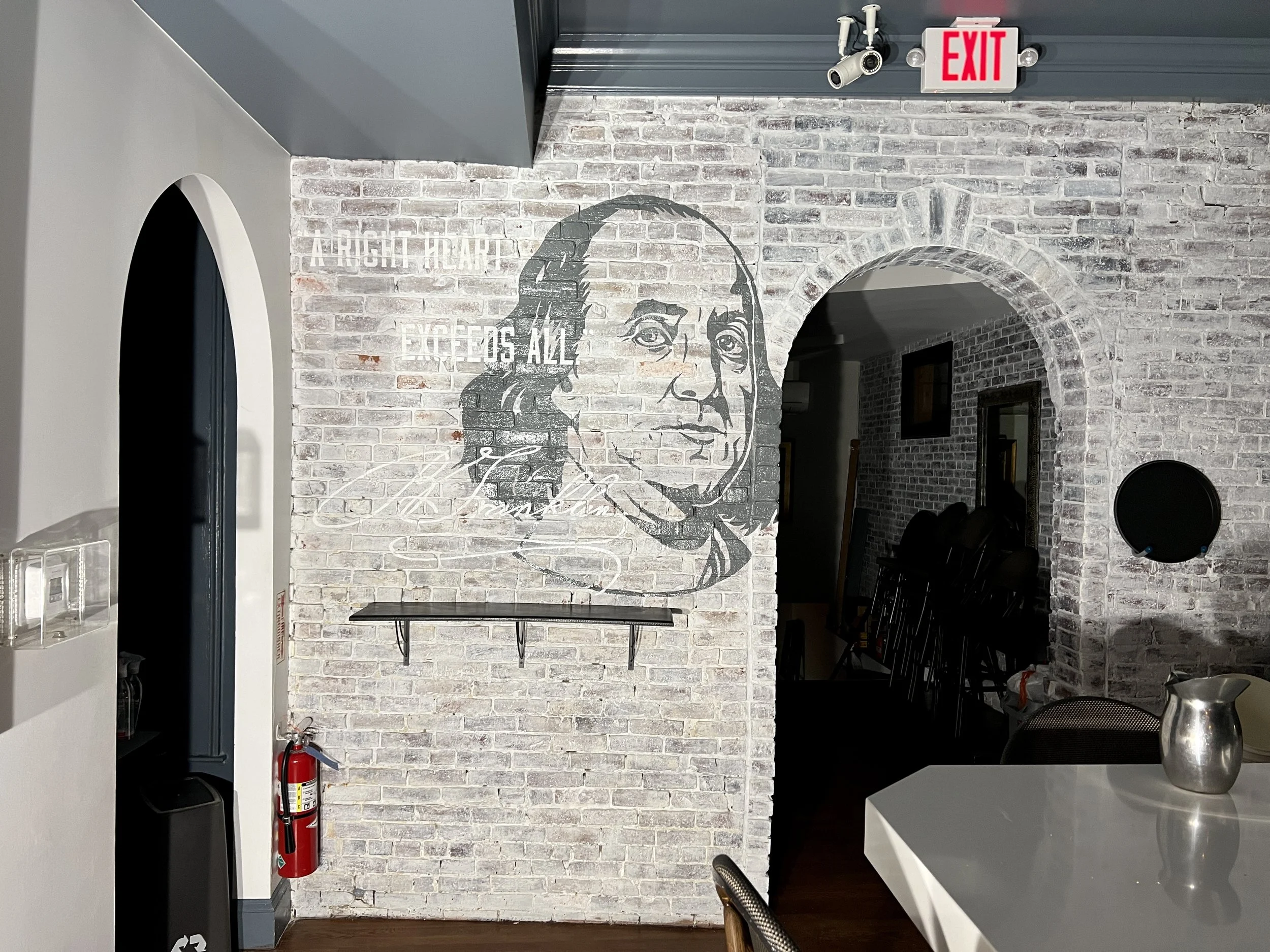Brick-walled restaurant interior with a large black-and-white portrait mural, arched doorway, exit sign, security camera, and dining table.